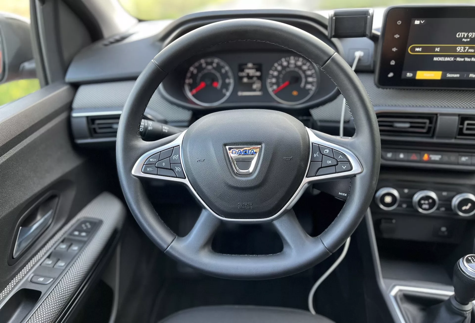 Vista interior desde el asiento del conductor de un Dacia Jogger 2022 mostrando el volante multifunción forrado en cuero, cuadro de instrumentos analógico con pantalla digital central, pantalla de infoentretenimiento, control climático y palanca de cambios manual