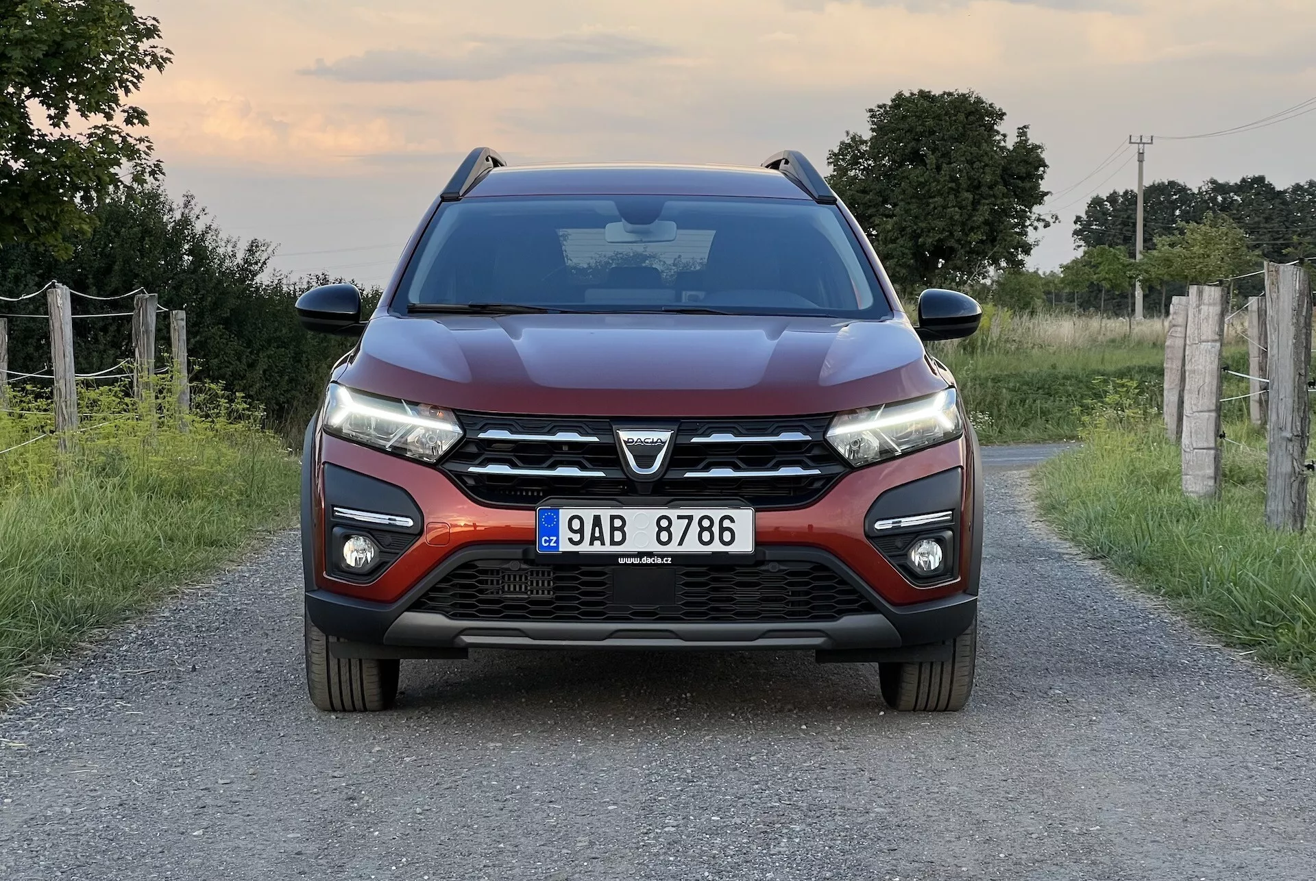 Dacia Jogger 2022 posicionado en una estrecha carretera rural, presentando una vista frontal simétrica que destaca el diseño moderno del coche, incluyendo una parrilla frontal prominente con un gran emblema, elegantes faros LED, líneas contorneadas en el capó, parachoques frontal con luces antiniebla y sensores de aparcamiento, y espejos laterales con señales de giro integradas.