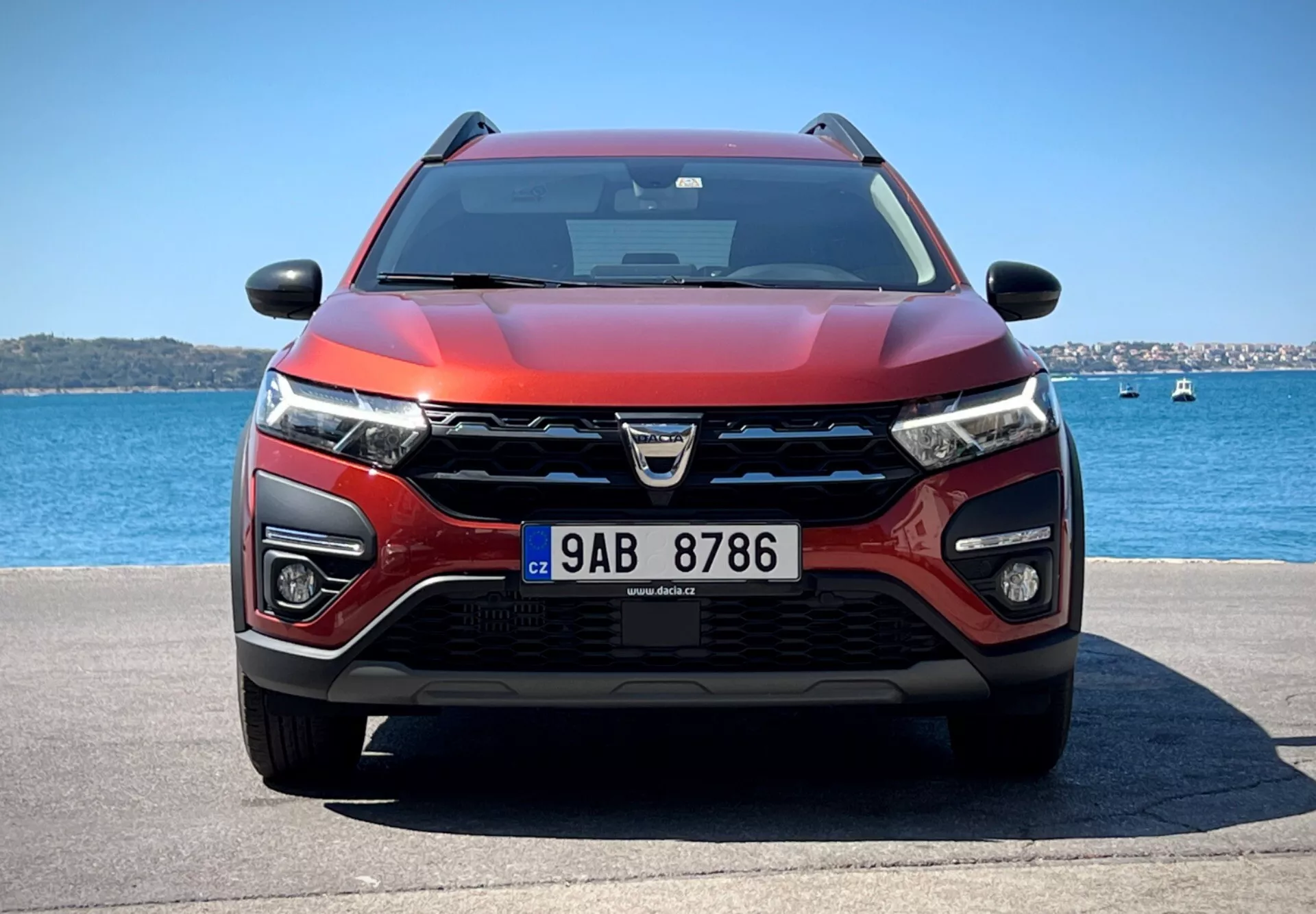 Dacia Jogger 2022 mostrando su diseño moderno de crossover y avanzadas características tecnológicas en un sereno entorno acuático, con una parrilla prominente con logo, faros LED, luces antiniebla, posibles sensores ADAS, tomas de aire para refrigeración y radar, esquema de color rojo y negro de dos tonos, capó aerodinámico, equipado con limpiaparabrisas que indica posible tecnología sensible a la lluvia, espejos laterales con señales de giro integradas posiblemente conteniendo tecnología de monitoreo de punto ciego, bajo cielos despejados.