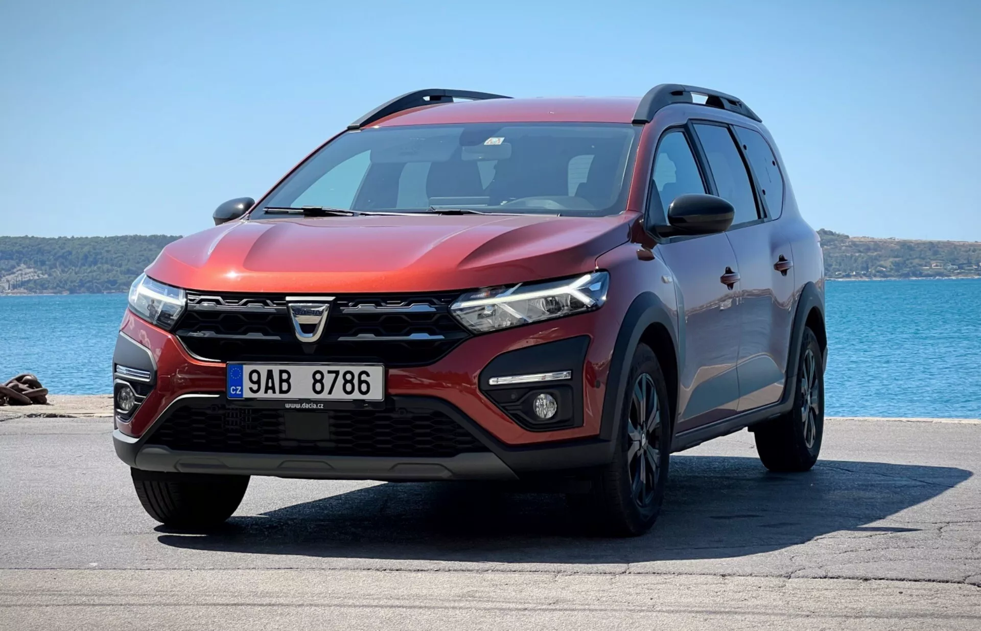 Vista frontal de tres cuartos del SUV Dacia Jogger 2022 estacionado con fondo oceánico, mostrando diseño de parrilla distintivo, faros LED, luces de niebla, parachoques delantero aerodinámico con entradas de aire y sensores de estacionamiento, contornos musculosos del capó, ruedas de aleación, señales de giro integradas en los espejos laterales, perfil lateral con revestimiento protector, rieles de techo, ventanas tintadas y exterior rojo metálico bajo la luz solar brillante indicando perfil de diseño, características tecnológicas y utilidad.