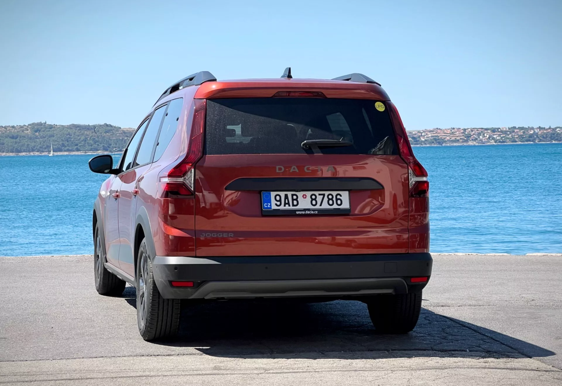Vista trasera de un SUV compacto rojo Dacia Jogger 2022 estacionado en un área pavimentada frente al agua bajo la luz brillante del día, mostrando la parte trasera completa del vehículo incluyendo las luces traseras, la ventana trasera y la placa de matrícula contra un fondo de agua azul y cielos despejados.