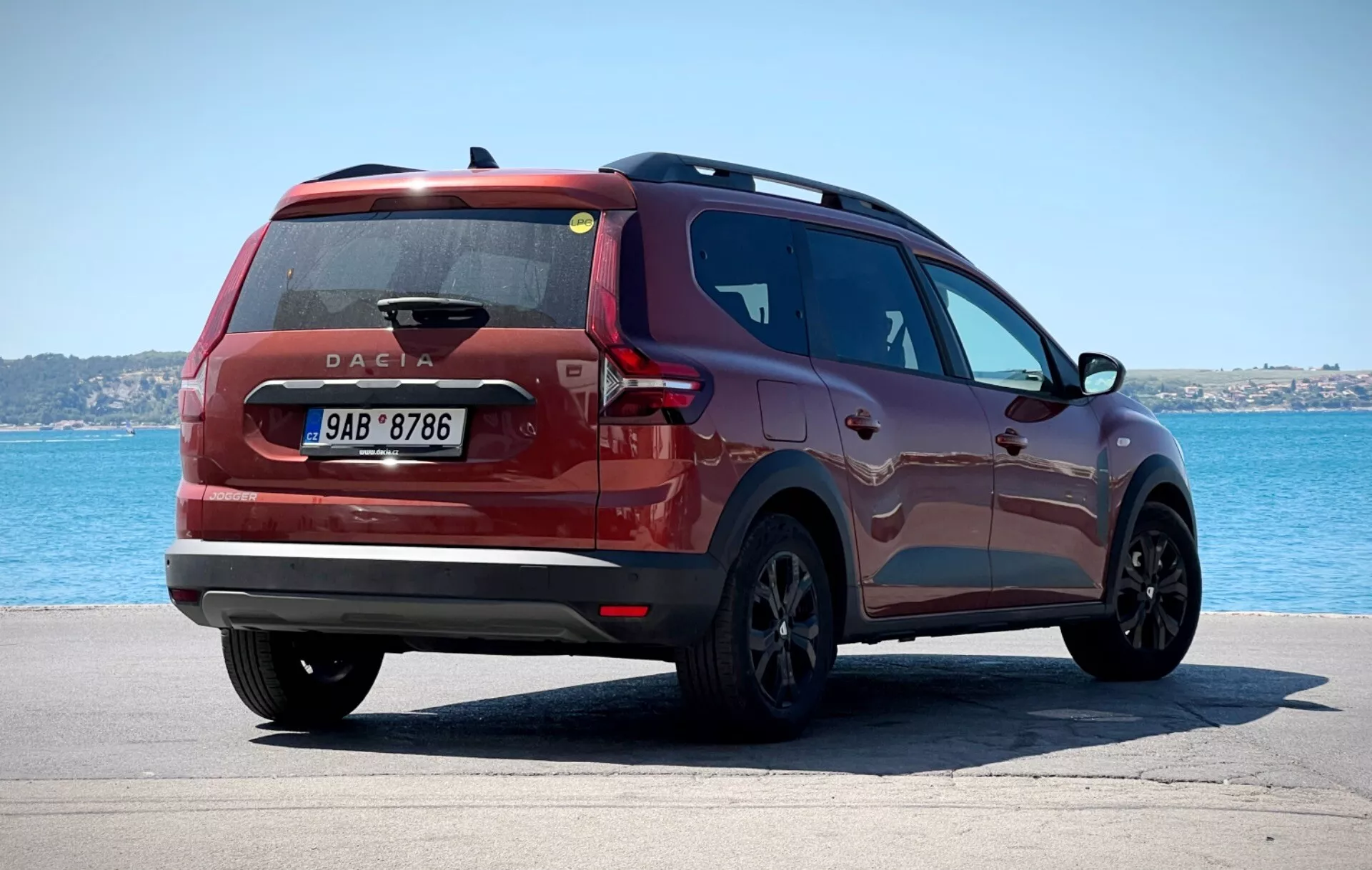 Dacia Jogger 2022 fotografiado en una vista trasera de tres cuartos posicionado sobre una superficie pavimentada con un cuerpo de agua y cielo despejado en el fondo, mostrando luces traseras modernas, un distintivo de Dacia colocado centralmente y un perfil lateral elegante contra un tranquilo entorno costero o lacustre.