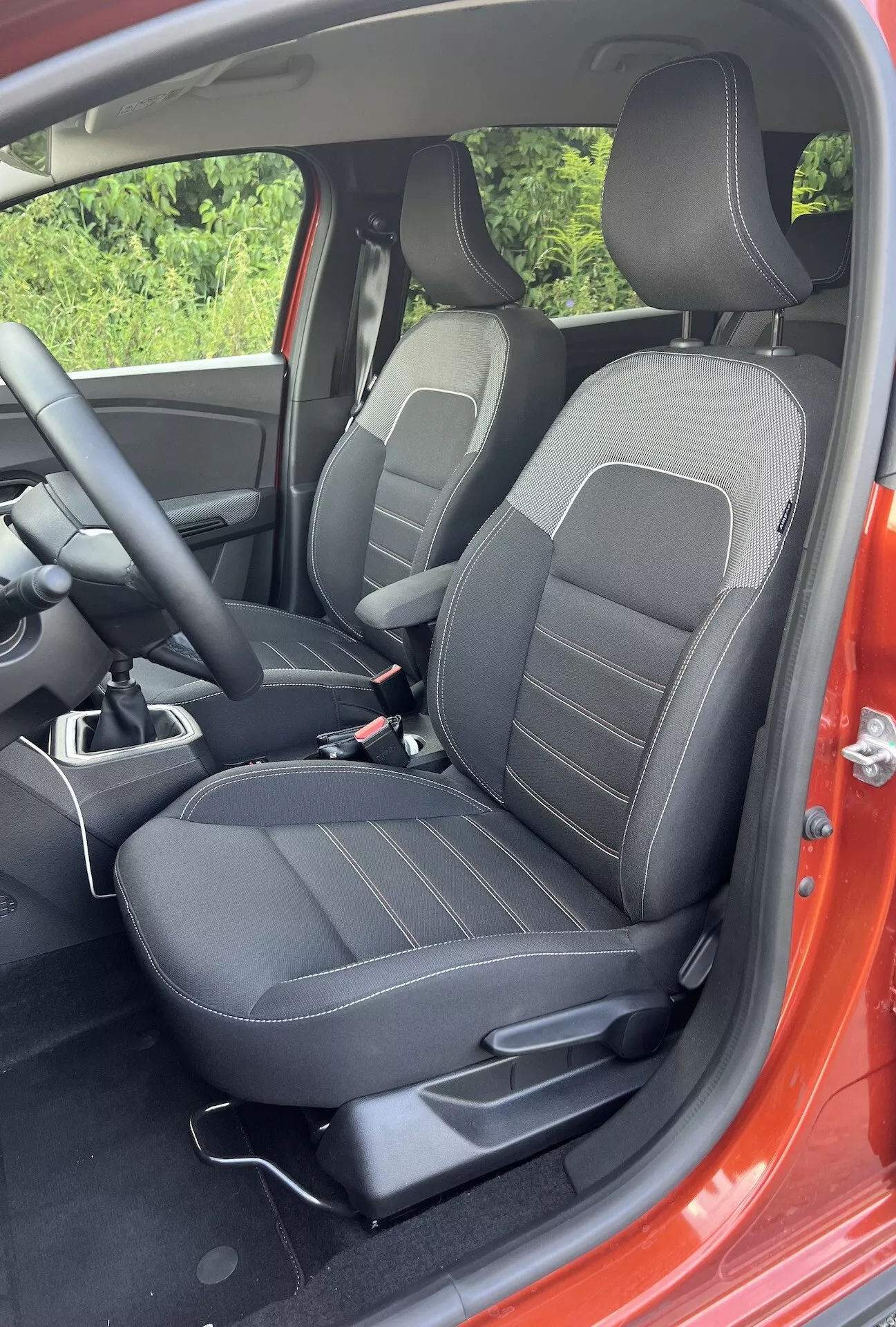 Vista detallada del interior del Dacia Jogger 2022 que muestra el área del pasajero delantero con asientos tapizados en negro con patrones de líneas, controles de ajuste, parte de la consola central y un atisbo del exterior rojo