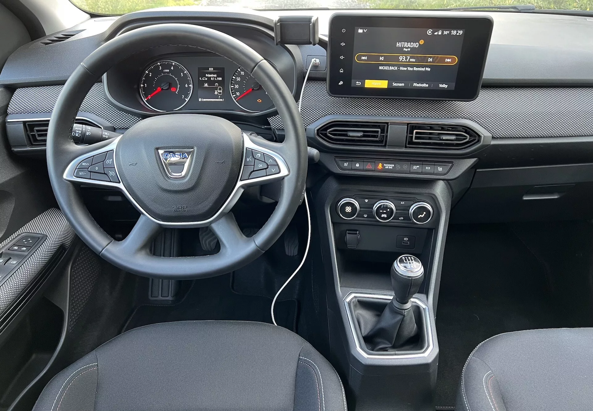 Vista del interior de un Dacia Jogger 2022 desde el asiento trasero, destacando el tablero del conductor, volante con logo del fabricante y botones de control, cuadro de instrumentos digital-analógico, sistema de infoentretenimiento táctil, controles HVAC, control climático de dos zonas y palanca de cambios manual