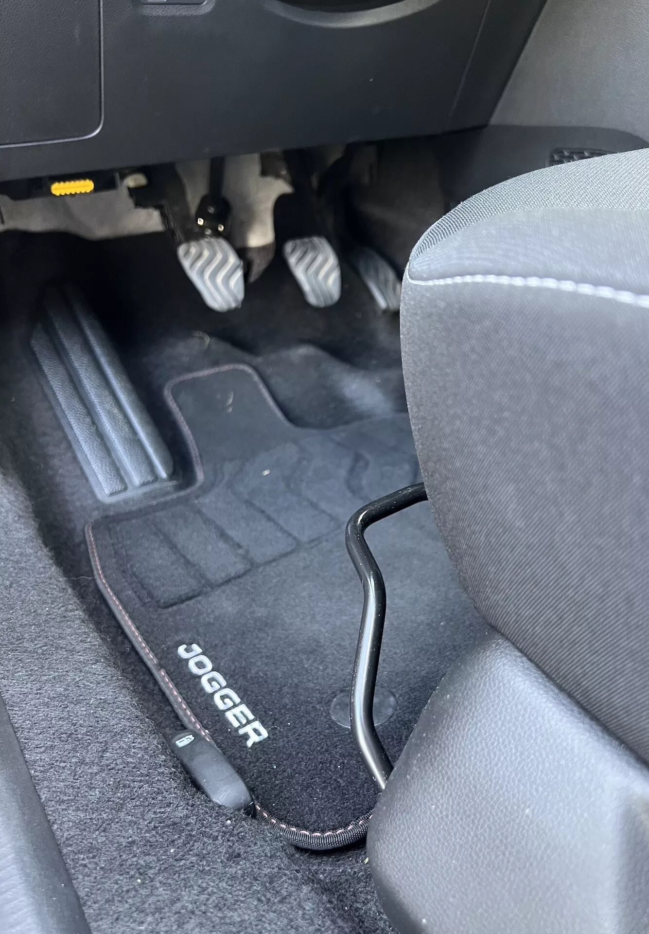 Interior del Dacia Jogger 2022 mostrando el espacio para los pies del lado del conductor con pedales metálicos, tapete de piso de marca, asiento de tela gris y ribete de plástico negro