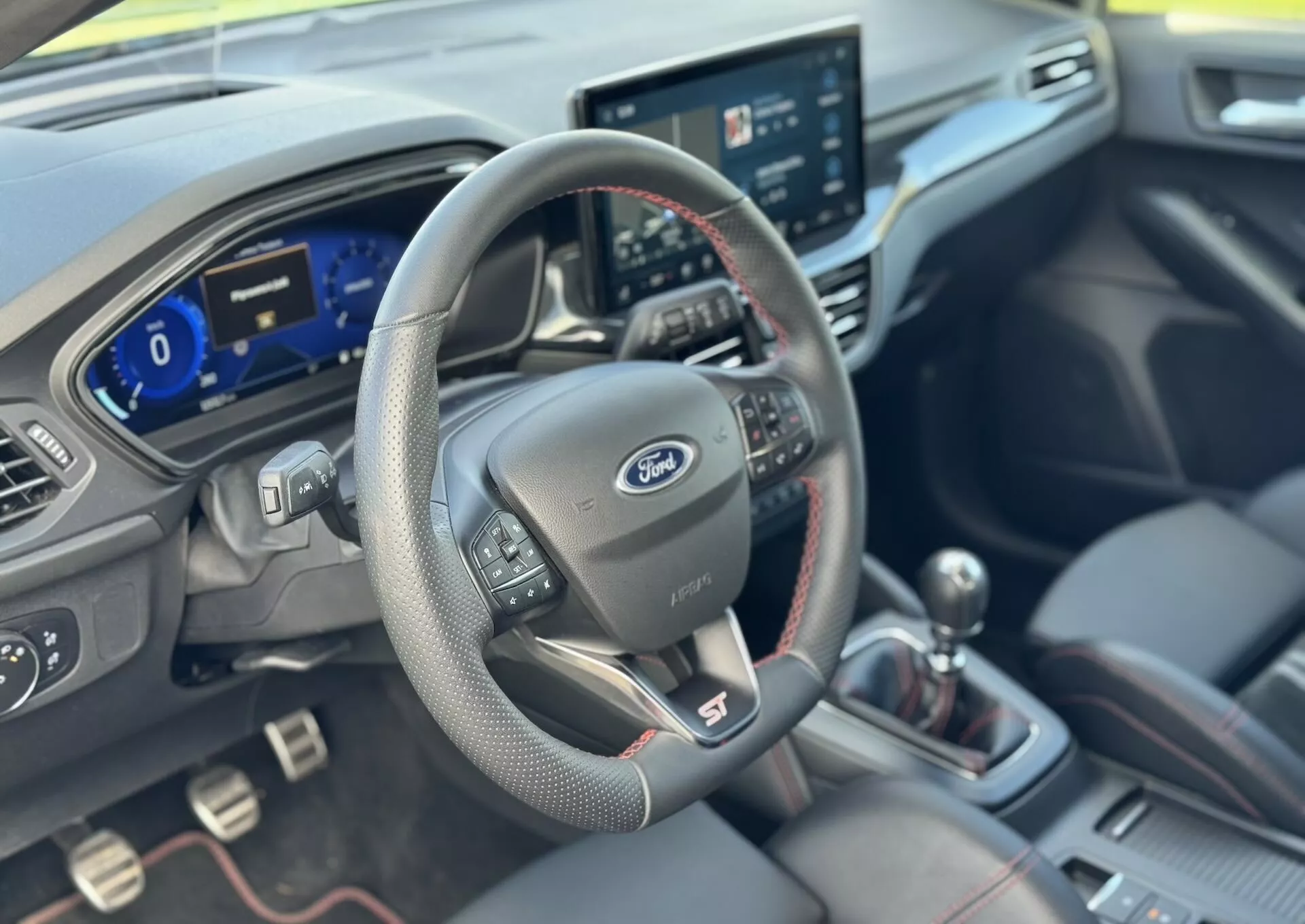 Interior del Ford Focus ST 2022 mostrando la perspectiva del conductor con volante cubierto de cuero con controles multimedia y de crucero, panel de instrumentos digital, sistema de infoentretenimiento táctil central, palanca de cambios manual con costuras rojas y asientos tapizados premium