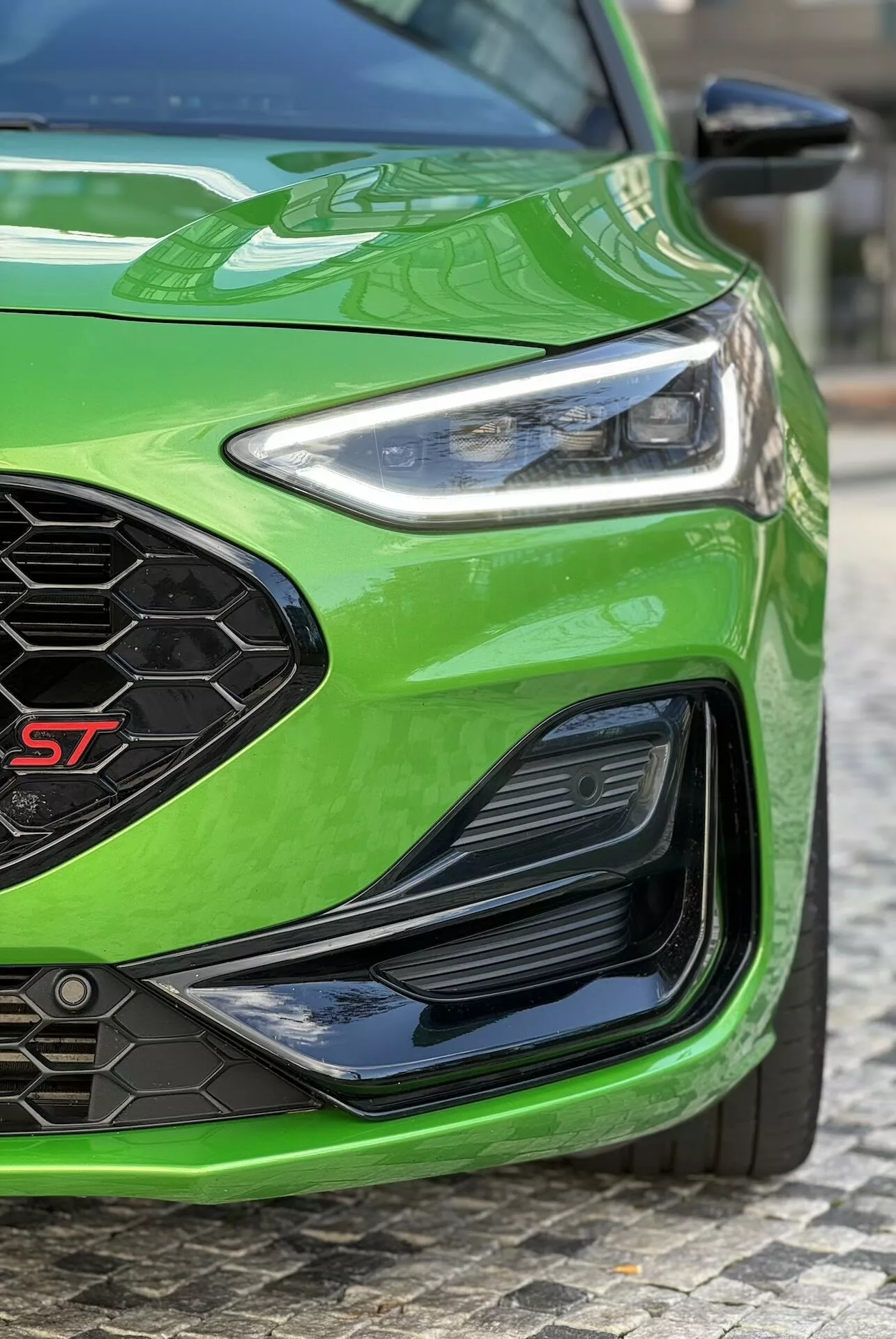 Vista angular en detalle de la sección delantera izquierda de un Ford Focus ST 2022 en verde vibrante, mostrando el faro LED, la rejilla frontal de panal con insignia ST, el parachoques inferior con entradas de aire y el acabado de pintura brillante, reflejando características avanzadas de iluminación, aerodinámica y rendimiento.