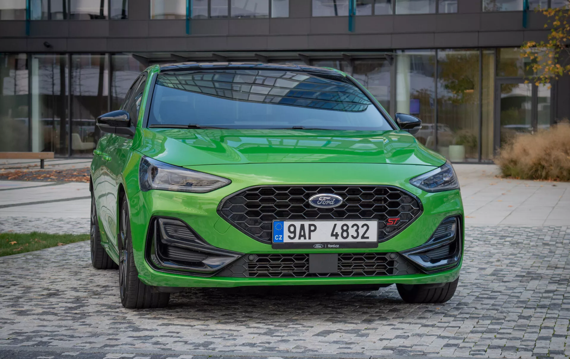 Ford Focus ST 2022 en verde vibrante, mostrado desde un ángulo frontal izquierdo, con una gran parrilla de panal de abejas, faros LED elegantes, parachoques delantero aerodinámico con spoiler y situado contra un telón de fondo urbano moderno