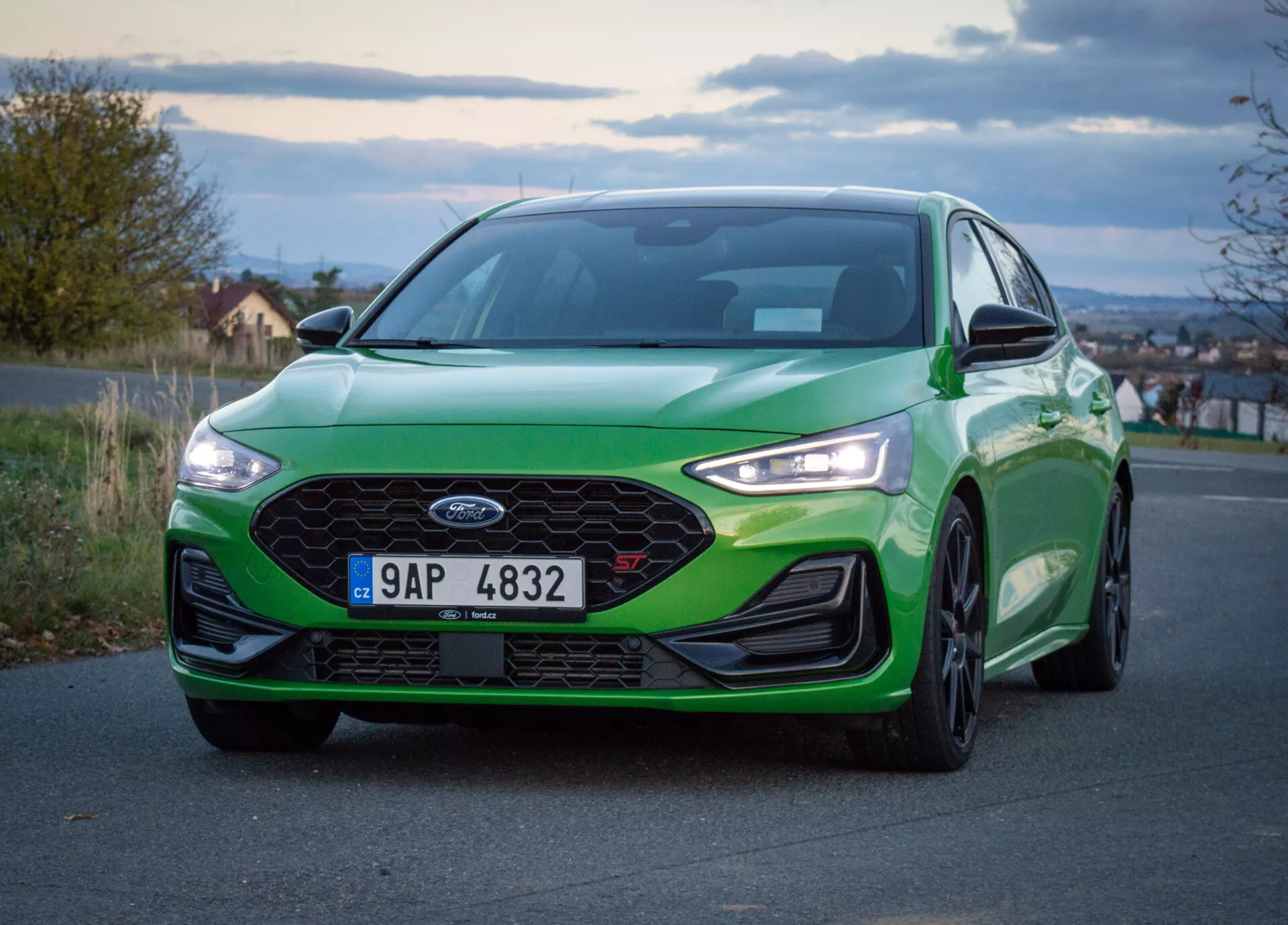 Ford Focus ST 2022, vista frontal en ángulo bajo, pintura verde con brillo vibrante, gran parrilla de panal, faros LED y parachoques delantero aerodinámico, en la carretera al amanecer o al anochecer