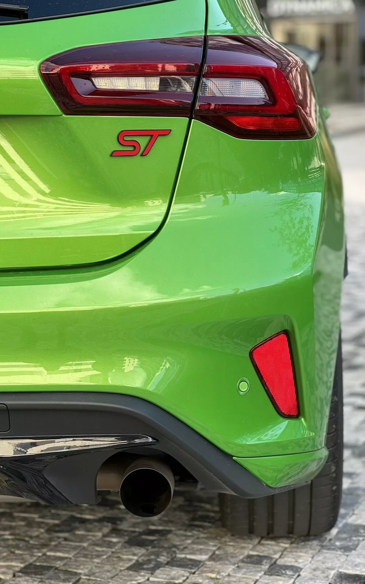Ford Focus ST 2022 en verde vibrante con insignia ST, vista parcial de luz trasera roja y clara, punta de escape cromada y elementos de diseño trasero deportivo sobre superficie de adoquines