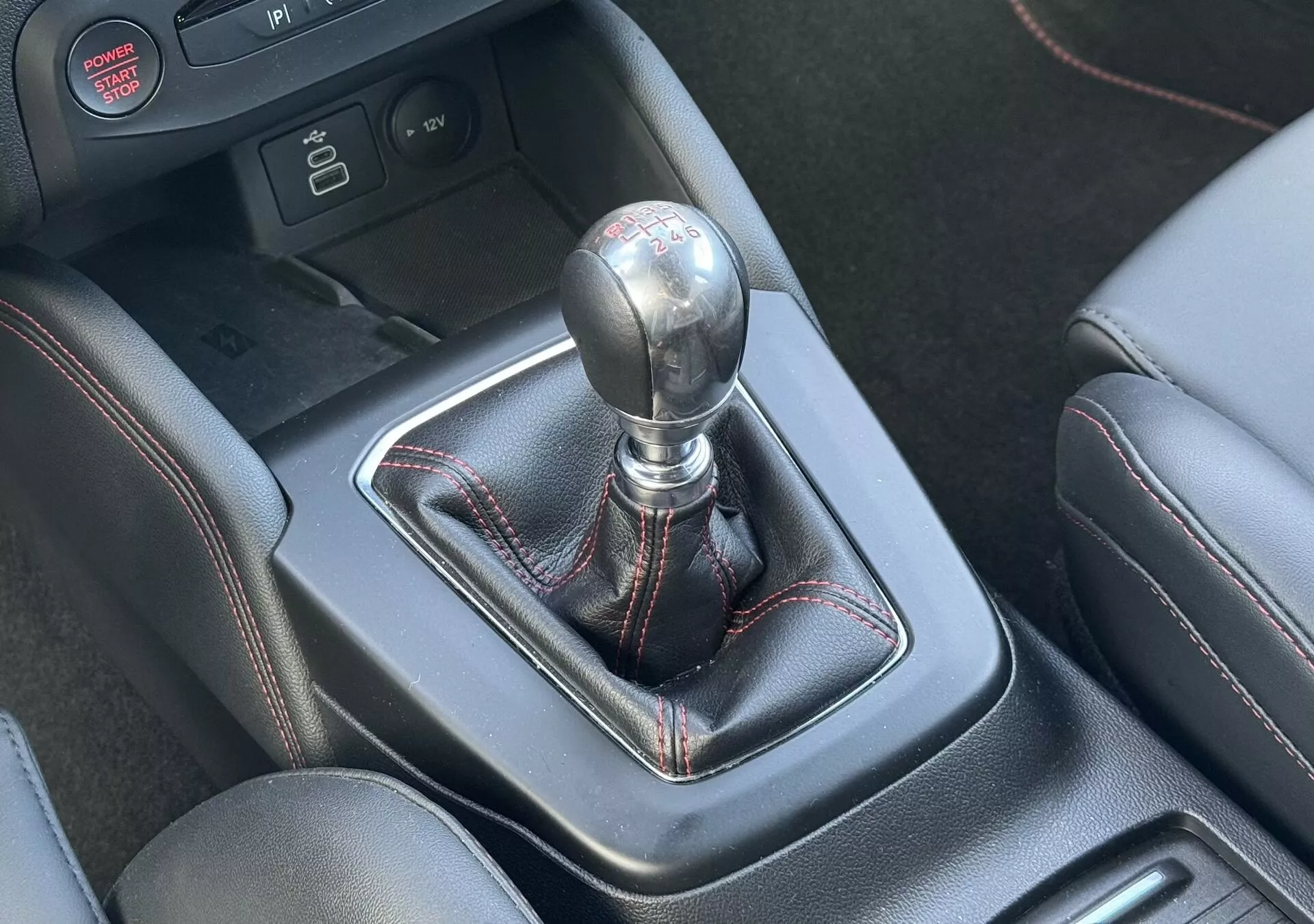 Primer plano del cambio de marchas manual del Ford Focus ST 2022 con pomo metálico, funda de cuero con costuras rojas y consola central que presenta ayudas a la conducción de alta tecnología