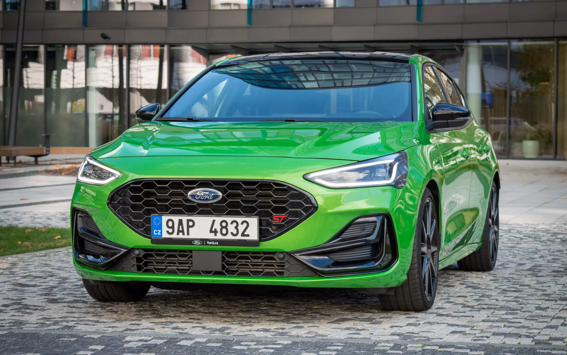 Ford Focus ST 2022 en verde vibrante, mostrando faros LED, parrilla de panal y diseño aerodinámico desde un ángulo dinámico bajo frontal tres cuartos con fondo urbano moderno
