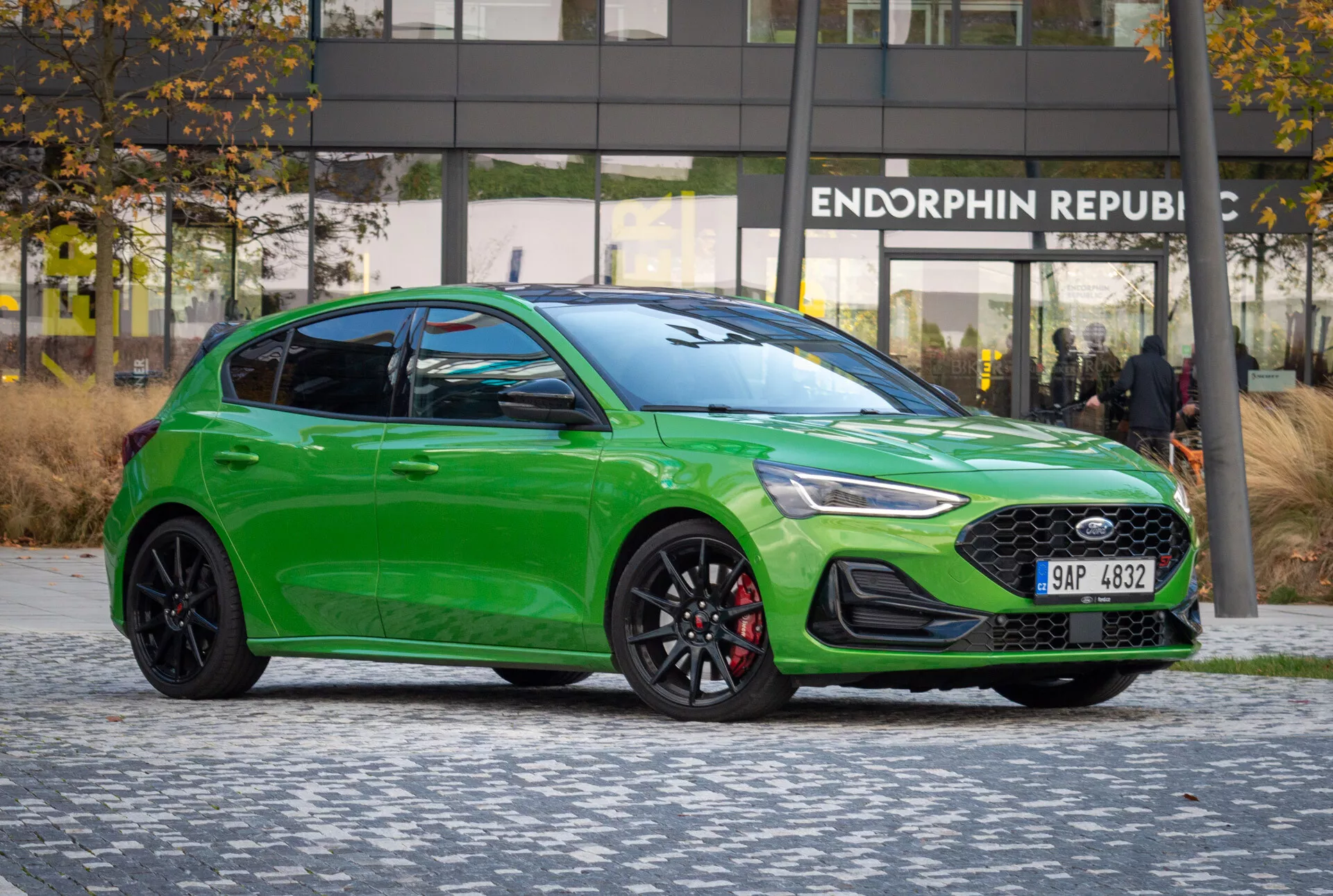 Ford Focus ST 2022 en verde vibrante estacionado en pavimento estampado, mostrando su perfil deportivo lateral-frontal, líneas de diseño, detalles visibles de faros y parrilla, ruedas enfocadas en rendimiento y pinzas de freno, bajo una iluminación favorable sin reflejos contra un telón de fondo urbano con edificio de oficinas moderno