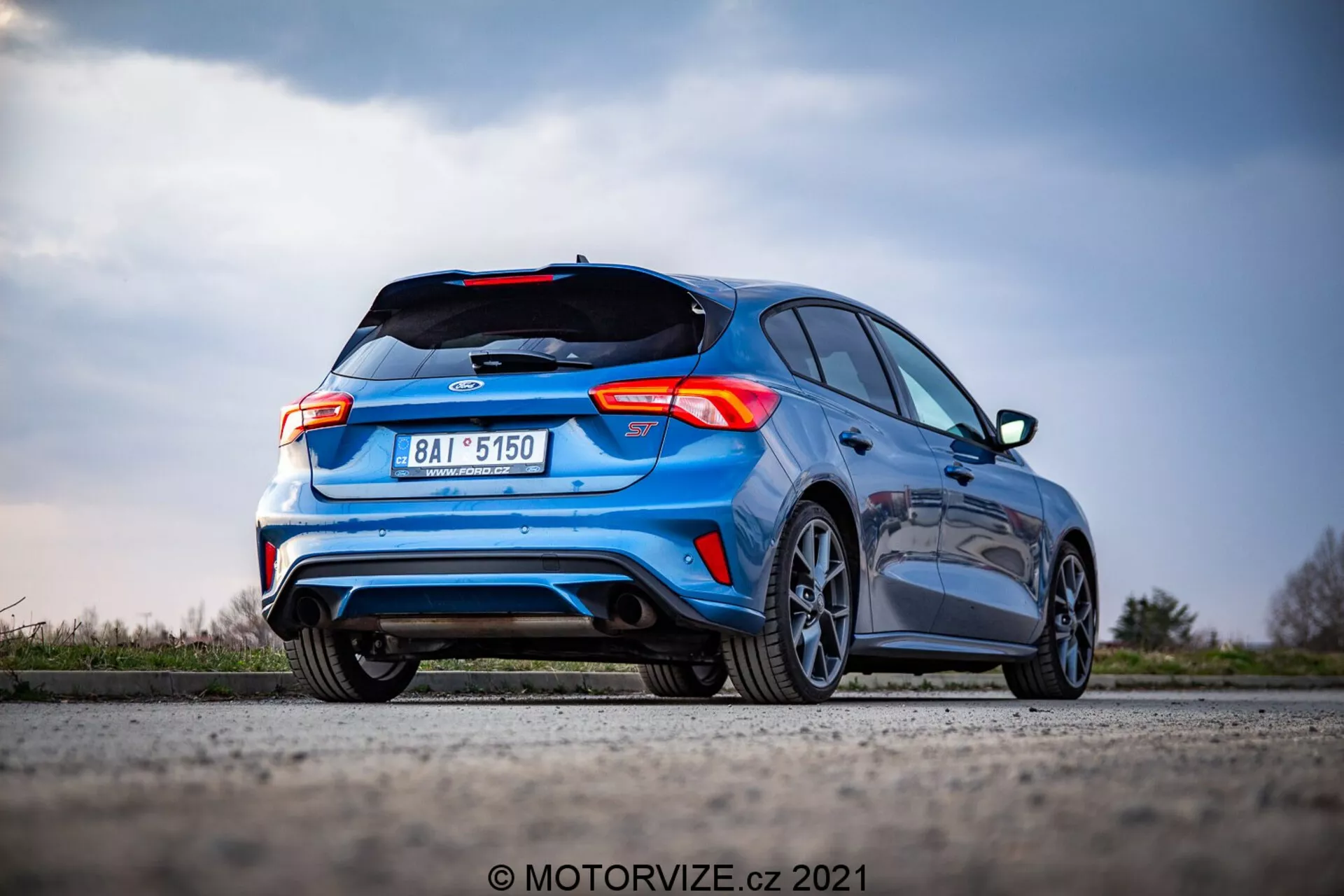 Imagen del Ford Focus 2022 desde un ángulo trasero bajo, mostrando su diseño hatchback, el emblema del fabricante en el maletero, diseño único de las luces traseras, parachoques trasero con difusor, dobles tubos de escape, alerón trasero, llantas de aleación, matrícula centrada, color azul vibrante, contra un fondo de paisaje natural borroso y cielo. El auto está estacionado en una superficie plana, capturado en enfoque nítido con efecto bokeh en el fondo.
