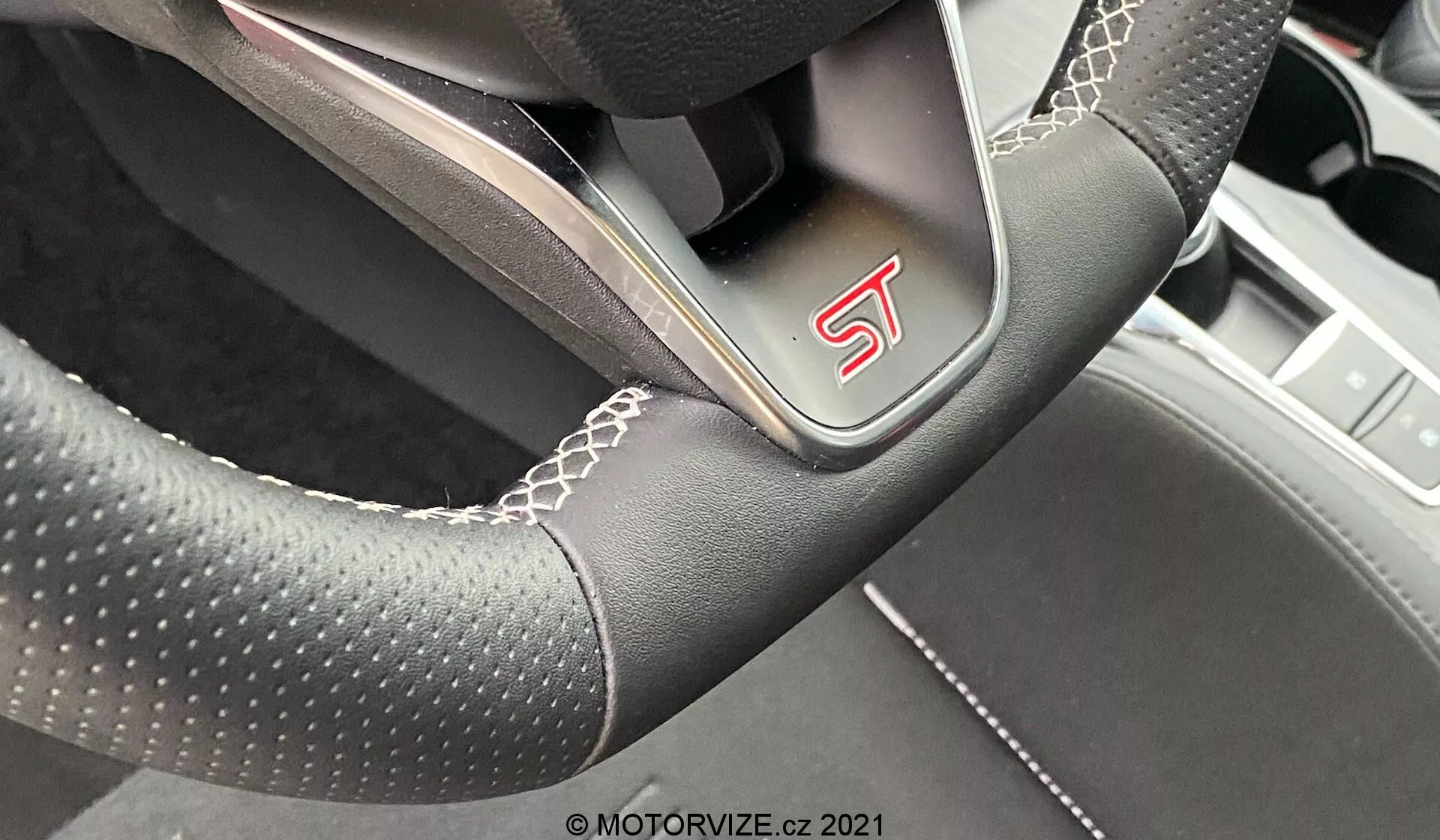 Vista de cerca de un volante moderno y deportivo desde la perspectiva del conductor en un Ford Focus 2022, con un emblema 'ST', material de cuero perforado, costuras blancas, diseño ligeramente aplanado en la parte inferior y radios metálicos que indican una artesanía orientada al rendimiento.