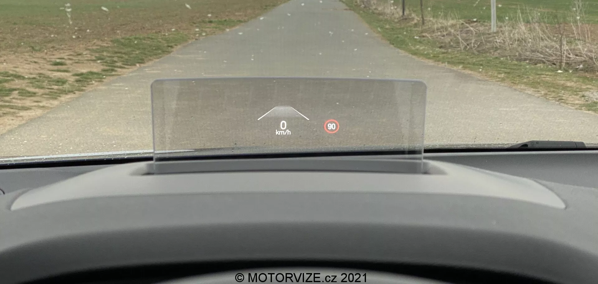 Vista desde el asiento del conductor de un Ford Focus 2022, mostrando el tablero curvado y minimalista y la pantalla de visualización frontal (HUD) prominente proyectada sobre el volante. La HUD muestra una velocidad de 0 km/h y un ícono de advertencia, sugiriendo que algo requiere la atención del conductor. El coche parece estar estacionado en una carretera rural o suburbana, con césped y árboles flanqueando la carretera, enfatizando un entorno tranquilo y no urbano.