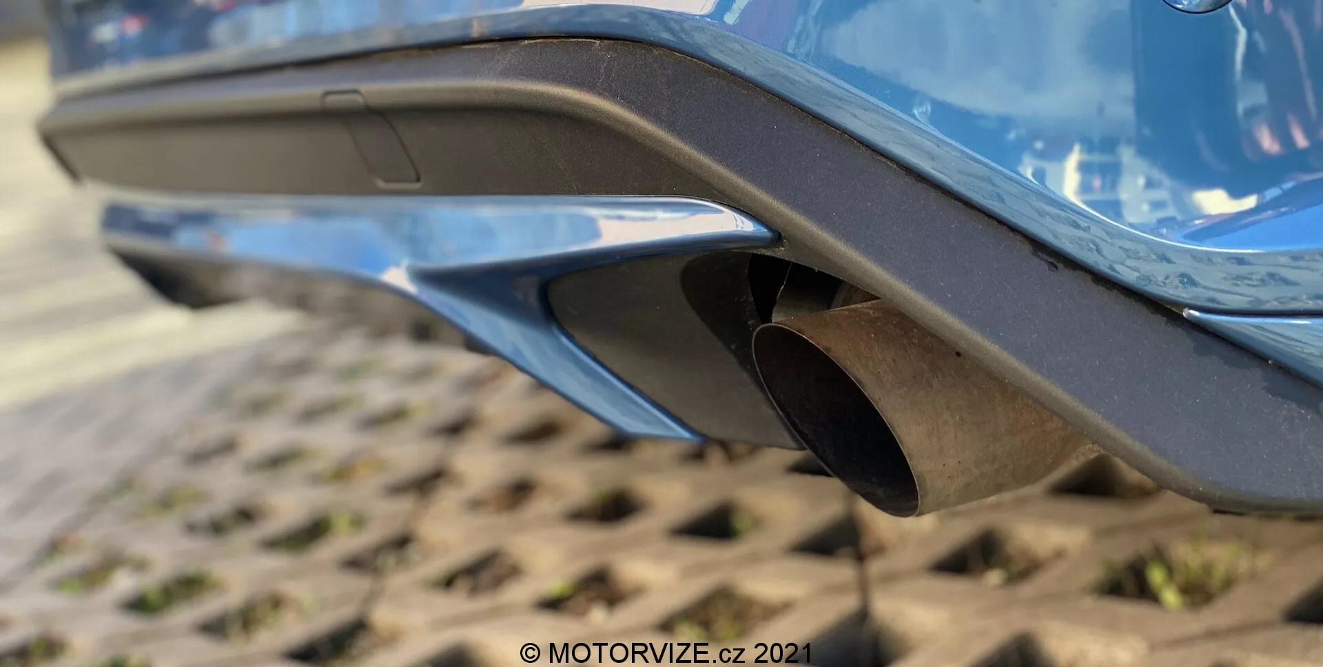 Primer plano de la sección trasera de un Ford Focus 2022, centrándose en el sistema de escape. La imagen, tomada desde un ángulo bajo, destaca la parte inferior del parachoques trasero con un diseño moderno y aerodinámico, pintura brillante y características integradas como ventilaciones o difusores. En el centro, una punta de escape circular de metal con un acabado ligeramente desgastado sobresale del parachoques. La superficie texturizada debajo sugiere un área de estacionamiento industrial o de uso intensivo, con un vistazo al chasis del vehículo visible.