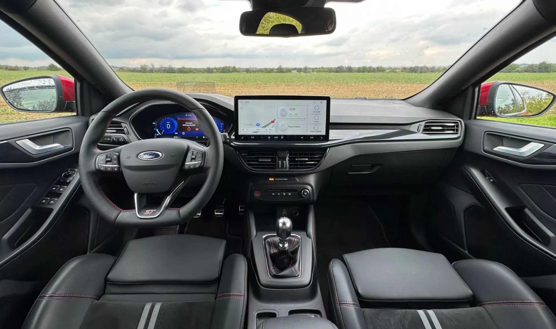 Vista interior del Ford Focus 2022 desde el asiento trasero mostrando el cuadro de instrumentos digital con velocímetro azul, volante con el logo de Ford, pantalla táctil de infoentretenimiento con navegación, selector de transmisión automática y asientos de cuero con costuras en contraste