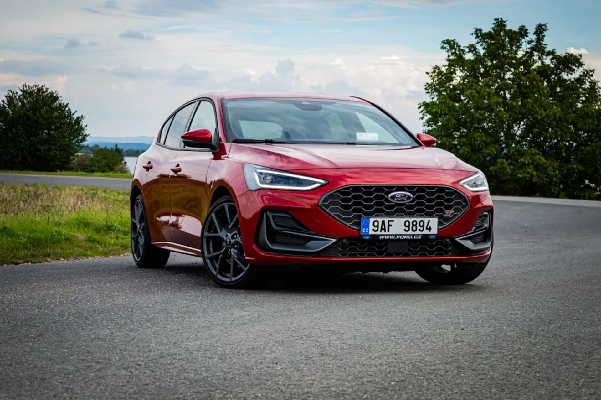 Hatchback rojo Ford Focus 2022 en carretera de asfalto, mostrando faros LED elegantes, llantas de aleación deportivas y un diseño moderno con líneas distintivas y una gran parrilla frontal contra un cielo nublado con toques de vegetación campestre