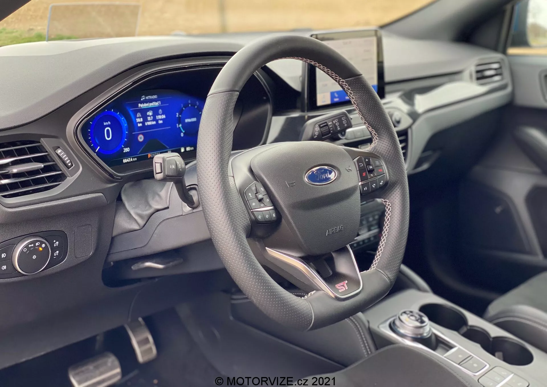 El interior de un moderno Ford Focus 2022, mostrado desde la perspectiva del asiento del conductor, destaca el volante con el logo de Ford, revestido en cuero con botones de control, y un panel de instrumentos totalmente digital que muestra el velocímetro y otras funciones. Los materiales de alta calidad y las tecnologías avanzadas, como la palanca de cambios automática, los controles de los faros y el sistema de infoentretenimiento central, indican un diseño sofisticado con interfaces amigables.
