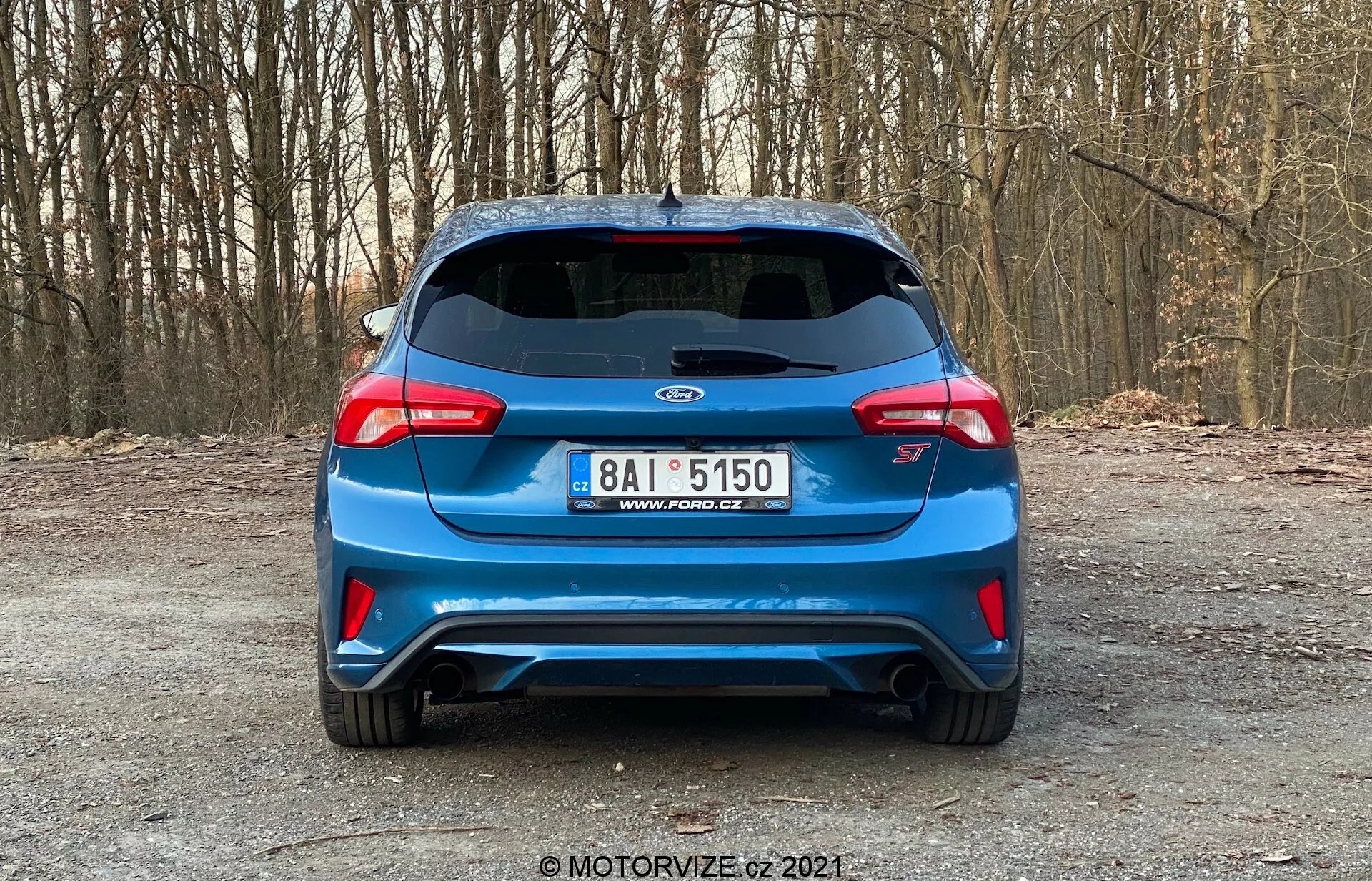 Vista trasera de un automóvil hatchback azul Kia 2022, posiblemente un modelo Proceed GT, estacionado en un camino no pavimentado en un bosque. La imagen muestra la tapa del maletero, el parachoques trasero y el espacio entre las distintivas luces traseras envolventes. Las características incluyen una placa de matrícula de diseño europeo centrada debajo del emblema del fabricante, el emblema 'Proceed GT' específico del modelo, un spoiler montado en el techo, limpiaparabrisas trasero, tercera luz de freno, doble salida de escape y un parachoques trasero de estilo agresivo con elementos ventilados. La foto clara y bien iluminada enfatiza el diseño deportivo del vehículo sin tecnologías avanzadas visibles, tomada desde un ángulo perpendicular estándar.