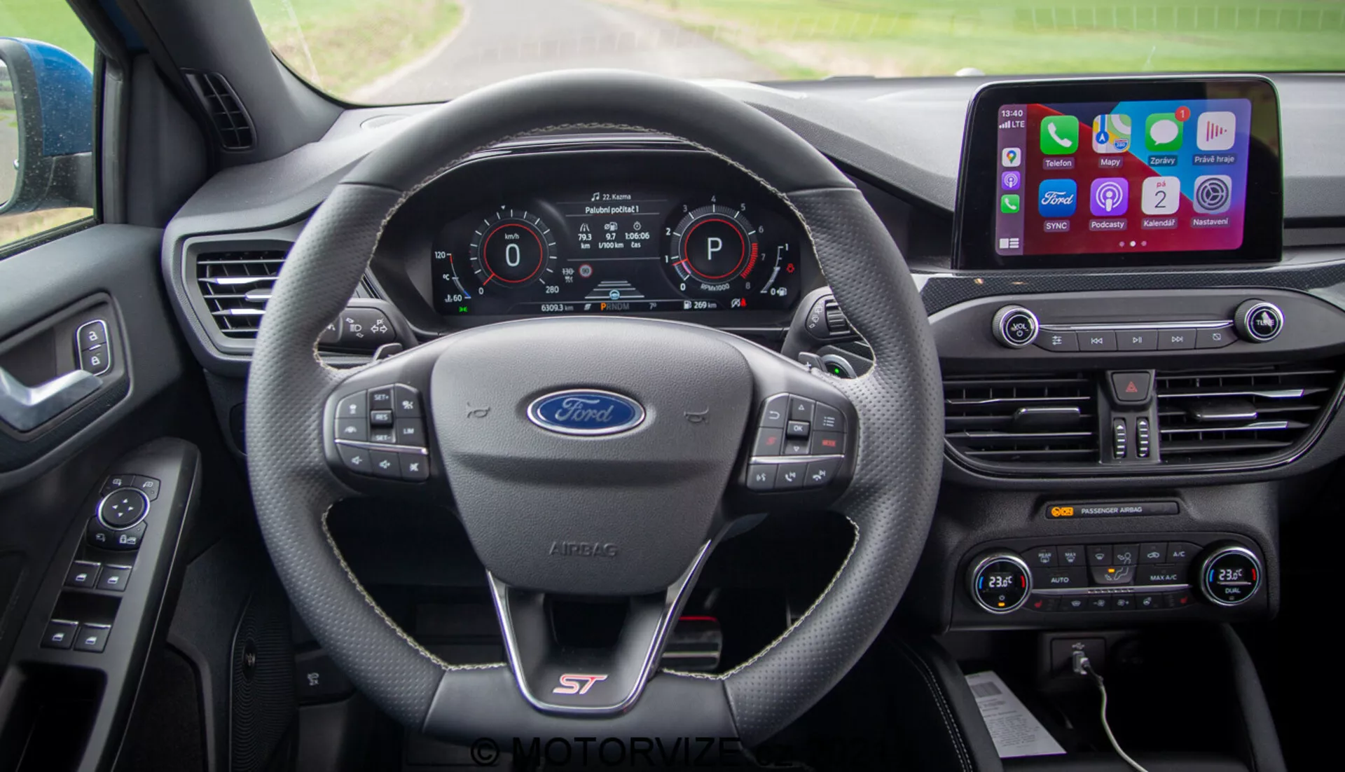 Vista interior del Ford Focus 2022 desde la perspectiva del conductor. Los elementos destacados incluyen un volante con el logotipo de Ford y el logo deportivo 'ST', un cuadro de instrumentos completamente digital que muestra velocidad, selección de marcha y estado de conectividad. La consola central alberga un sistema de infoentretenimiento con pantalla táctil grande con integración de smartphones, control de clima de doble zona y múltiples botones de control.