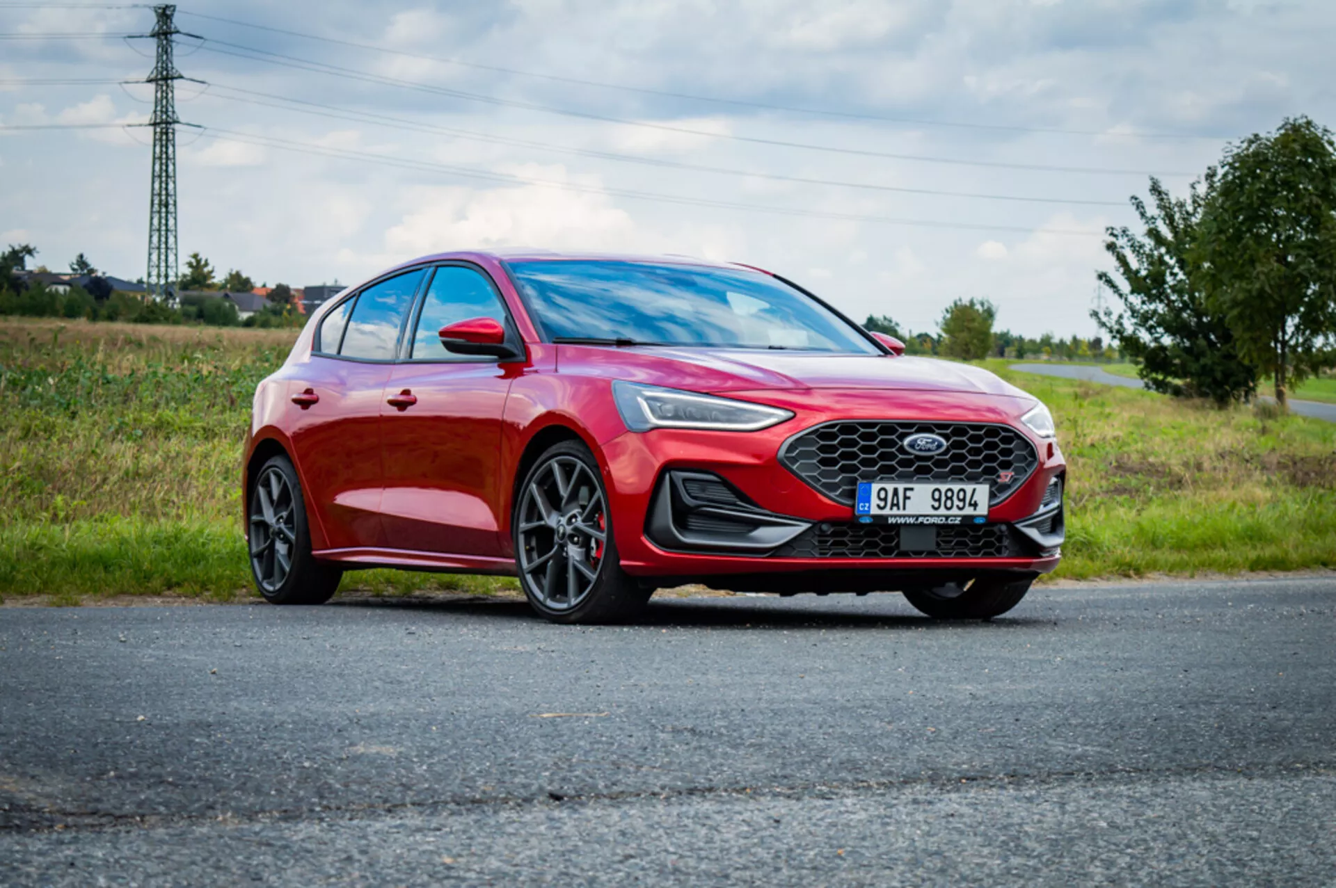Sedán moderno Ford Focus 2022 estacionado en carretera con cielo nublado y paisaje natural de fondo, destacando su color rojo metálico, rejilla frontal deportiva con emblema, faros delanteros LED angulares, parachoques frontal elegante y ruedas de aleación multirradio grandes desde una perspectiva frontal de tres cuartos en bajo ángulo