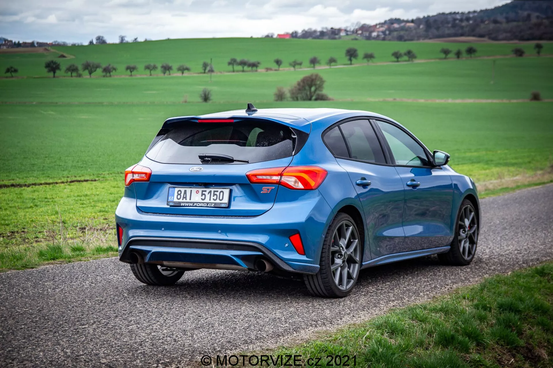 Vista tres cuartos trasera de un hatchback moderno azul, posicionado en un camino estrecho con un paisaje verde de fondo bajo un cielo nublado. La imagen captura el lado derecho y trasero del coche, destacando su alerón trasero deportivo, sistema de escape doble, llantas de aleación de múltiples radios y luces traseras distintivas. La postura del vehículo se realza con el ángulo bajo, enfatizando su aspecto dinámico. También se observan una insignia y una matrícula en la parte trasera.