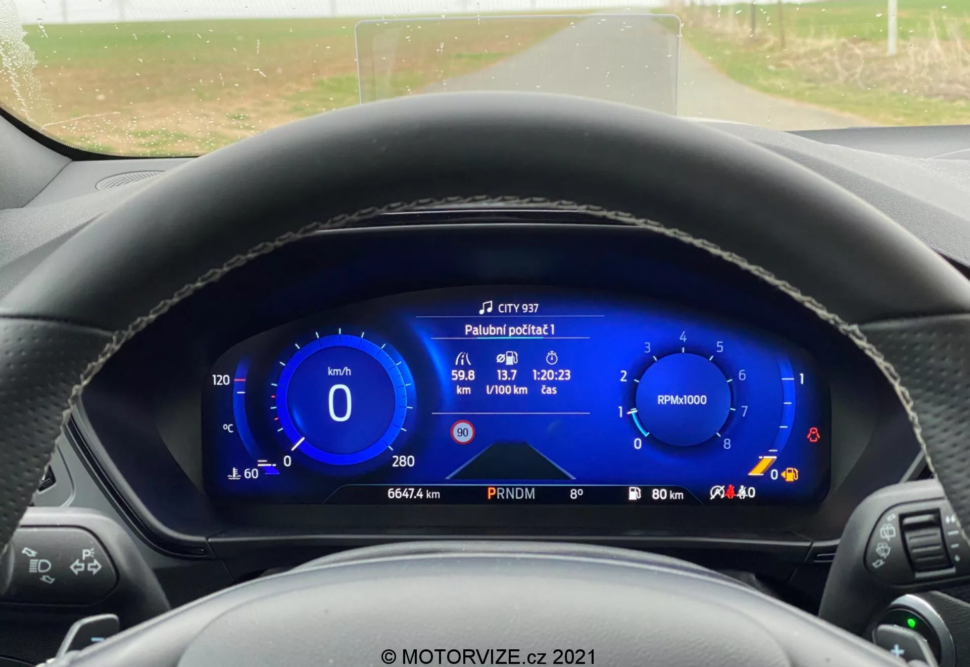 Vista en primera persona desde el asiento del conductor de un Ford Focus 2022, que muestra un panel de instrumentos totalmente digital. La pantalla digital, destacada en un vibrante color azul, incluye un velocímetro, un cuentarrevoluciones y diversa información del vehículo como el alcance de combustible, la temperatura exterior, la velocidad promedio, el tiempo de viaje y el consumo de combustible. El volante es parcialmente visible con palancas de control a ambos lados. A través del parabrisas, se ve una carretera rural de dos carriles en línea recta, flanqueada por árboles y arbustos bajo un cielo nublado.