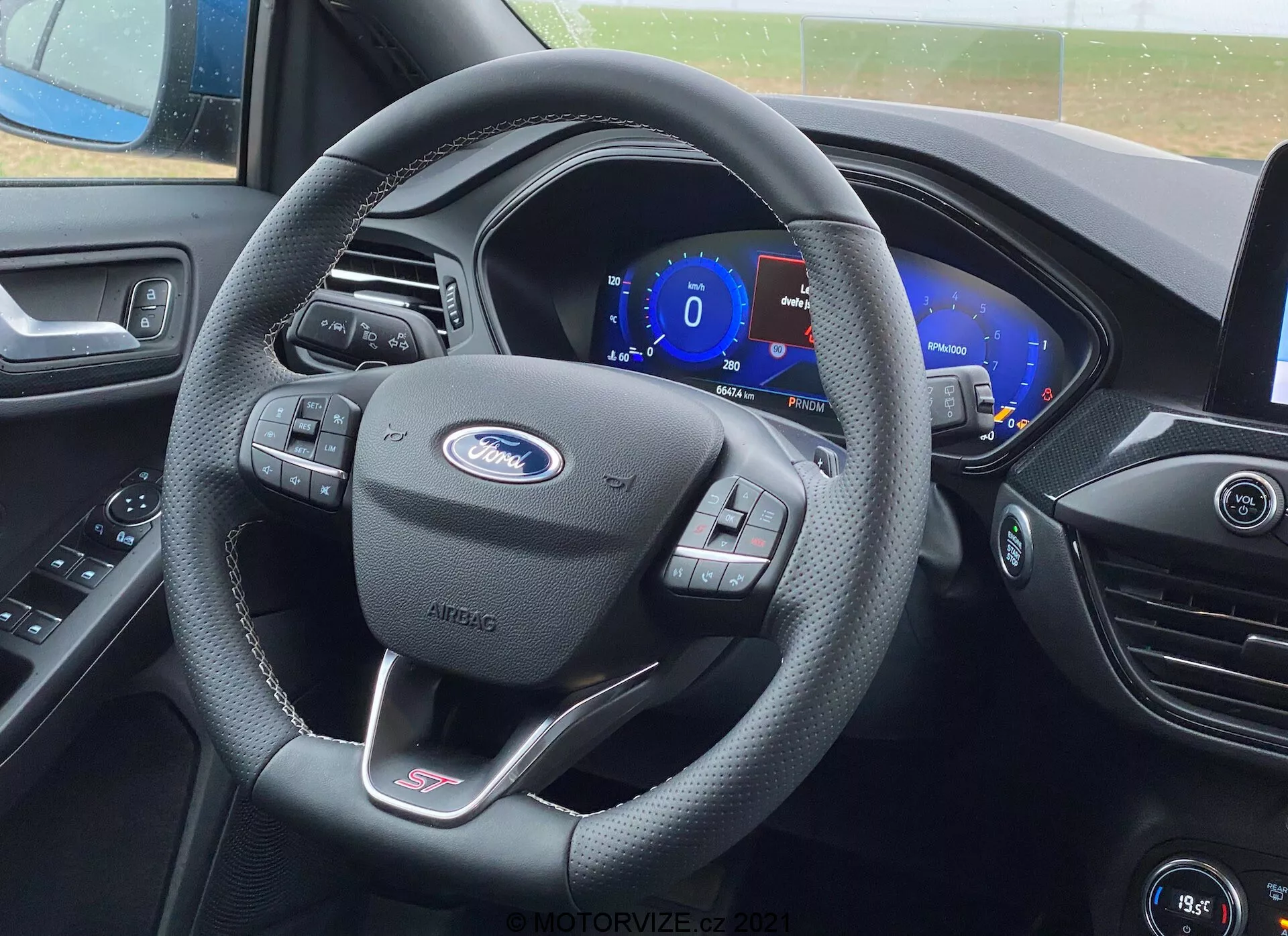 Vista clara del interior del asiento del conductor en un Ford Focus 2022, mostrando el volante envuelto en cuero negro con el prominente logo de Ford, insignia de la línea ST y controles multifuncionales. Detrás del volante hay un panel de instrumentos digital que muestra 0 mph. El tablero presenta contornos estéticamente agradables y una pantalla de infoentretenimiento y ventilaciones parcialmente visibles.