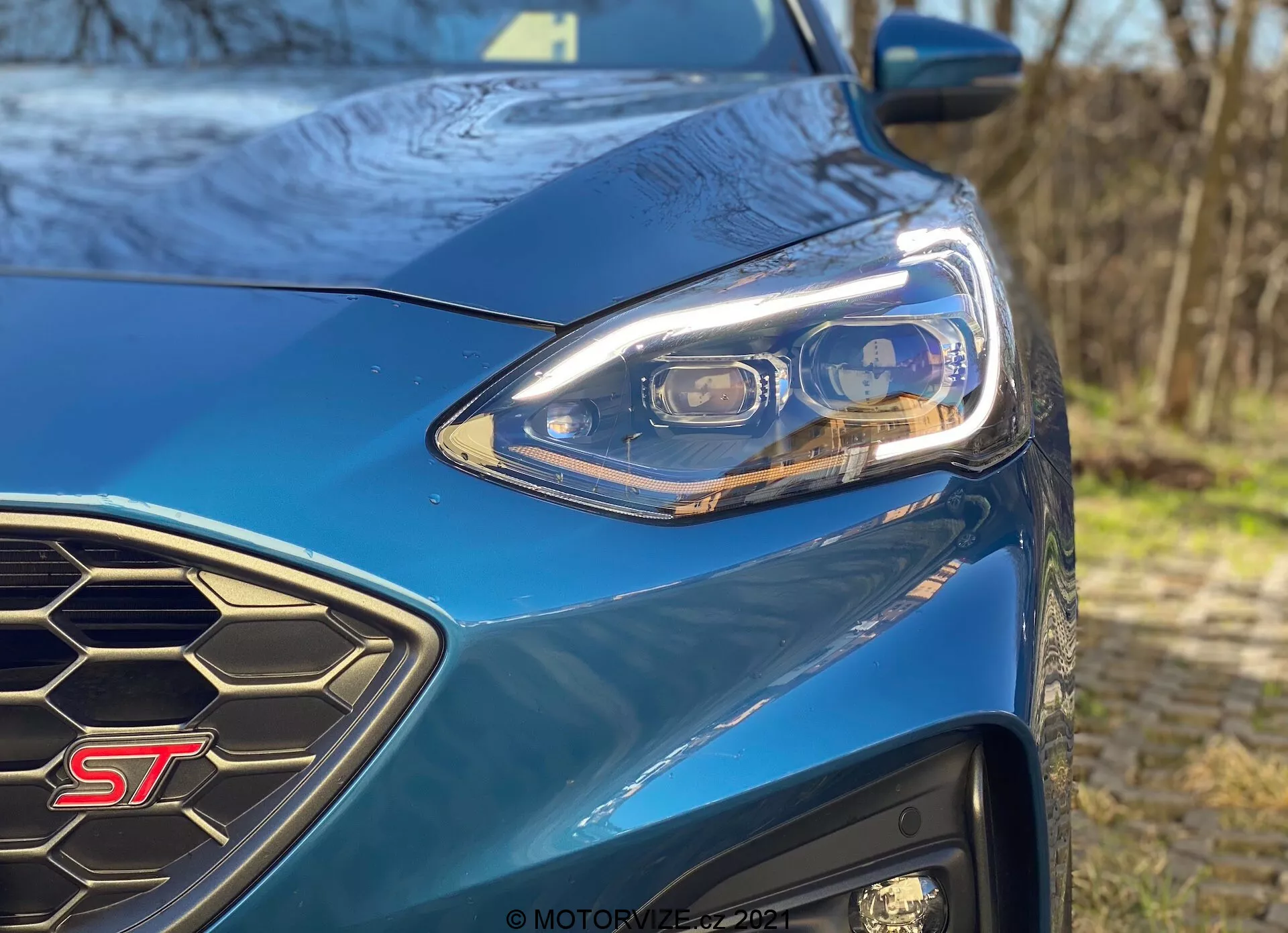 Primer plano del lado delantero izquierdo del modelo ST azul del Ford Focus 2022, que muestra el diseño moderno de los faros LED con múltiples elementos, la rejilla con patrón de panal y la insignia ST claramente visible en un entorno exterior bien iluminado.