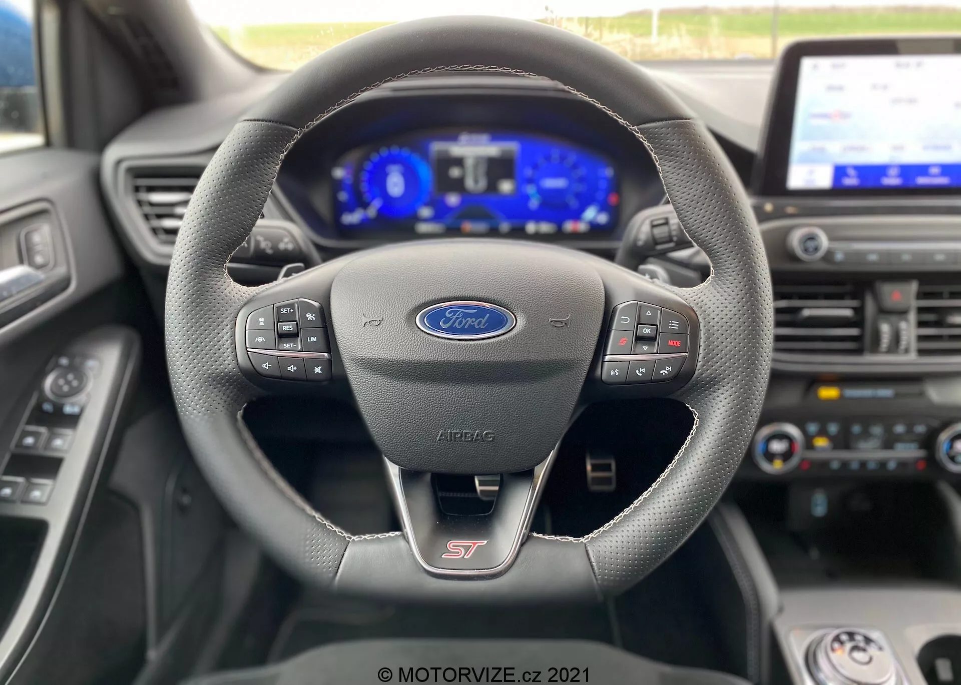 Vista interior del Ford Focus ST 2022, que muestra el volante deportivo con costuras de cuero con botones multifuncionales para medios y comandos de voz. El tablero de instrumentos completamente digital está iluminado, mostrando el velocímetro, el tacómetro y varios íconos de asistencia electrónica. Se ve una pantalla central de infoentretenimiento, que indica características tecnológicas avanzadas como navegación y sistemas de conectividad. La imagen resalta la interfaz moderna y tecnológica para el conductor desde la perspectiva del asiento del conductor, con la palanca de cambios en 'Parqueo'.