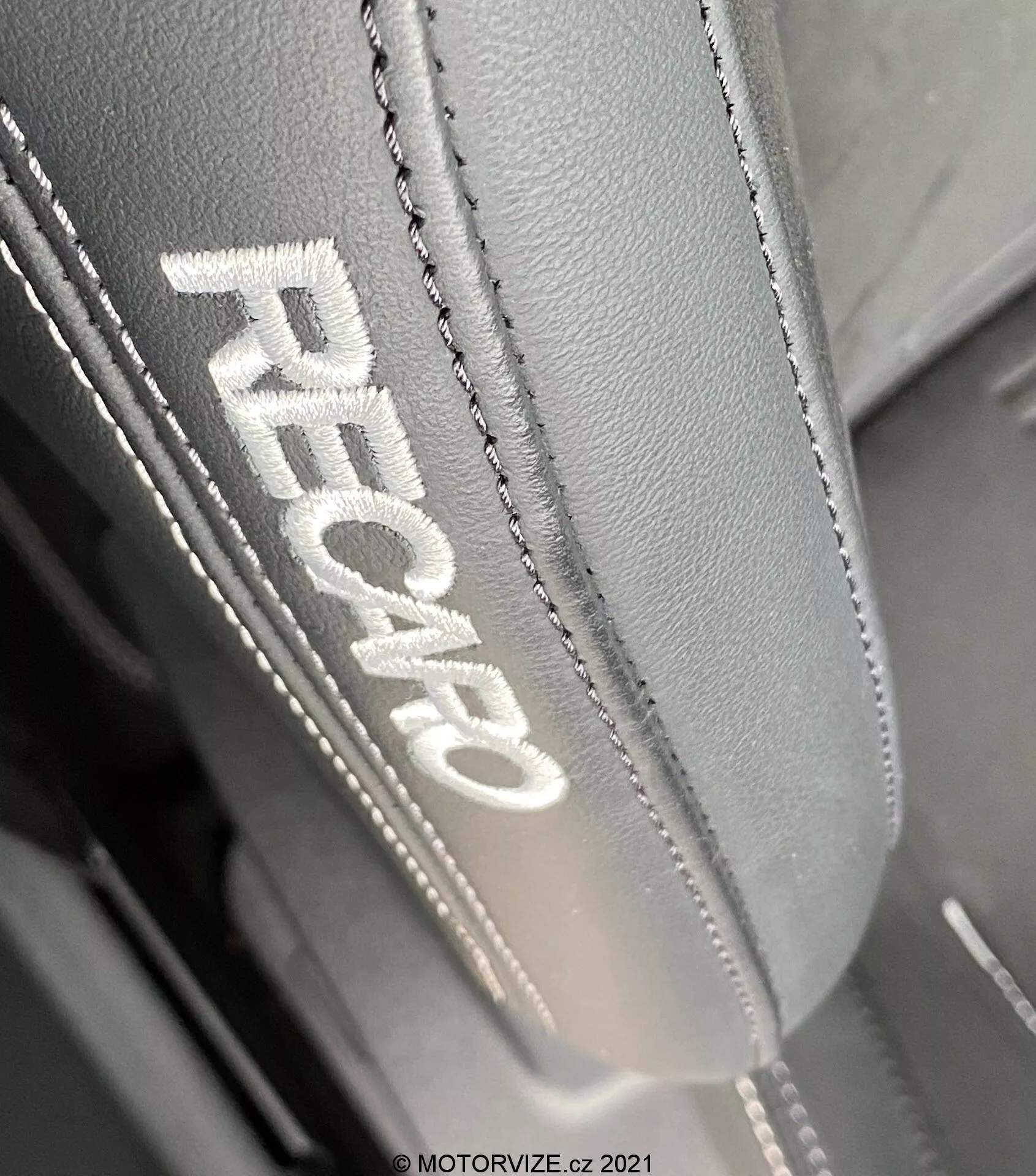 Vista de cerca del reposacabezas de un asiento de coche en un Ford Focus 2022, con bordado 'RECARO' indicativo de asientos de alto rendimiento. El lado izquierdo del reposacabezas se muestra prominentemente en una perspectiva sesgada de arriba hacia abajo, destacando el material fino de cuero o sintético de alta calidad con costuras detalladas y dobles. La iluminación revela la textura y calidad del tejido, enfatizando el diseño ergonómico destinado a proporcionar comodidad y soporte.