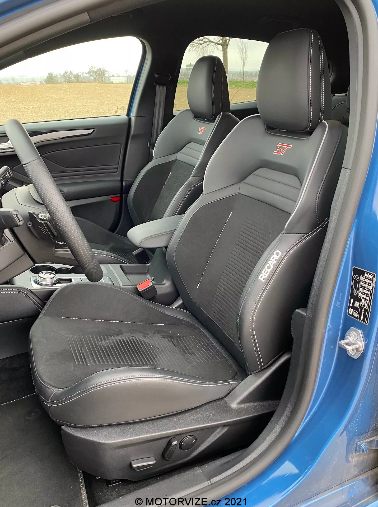 El interior de un Ford Focus 2022 se ve desde la perspectiva de la puerta del lado del conductor abierta, mostrando el asiento deportivo del conductor con las marcas 'ST' y 'RECARO'. El asiento está tapizado en cuero negro con tela en el centro para mejor agarre y comodidad, con un soporte lateral sustancial y reposacabezas integrado. También es visible el panel de la puerta en negro con un destello del color azul del exterior y los controles del asiento que insinúan ajuste eléctrico.