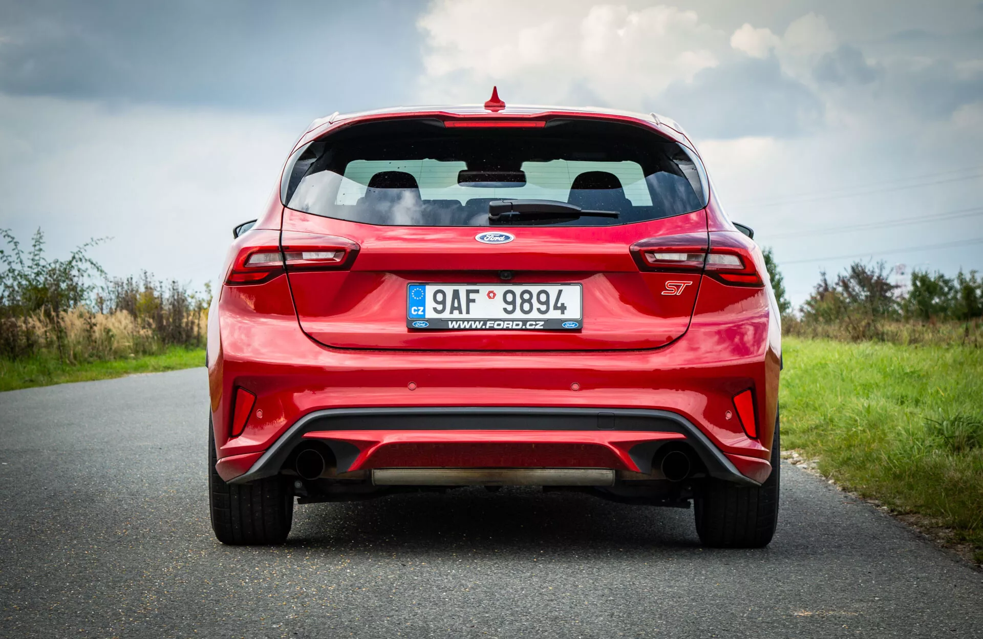 Ford Focus hatchback rojo 2022 capturado desde una perspectiva trasera y baja, mostrando su amplia postura, con luces traseras horizontales que envuelven los bordes, ventana trasera clara con limpiaparabrisas visible, un alerón deportivo arriba, y el distintivo del modelo del coche centrado en el maletero. También muestra una placa de matrícula, un parachoques deportivo con elemento difusor y ventilaciones de aire, puntas de escape dobles integradas en el parachoques, y luces reflectoras en los bordes del parachoques, situado contra una carretera y paisaje natural bajo un cielo nublado.