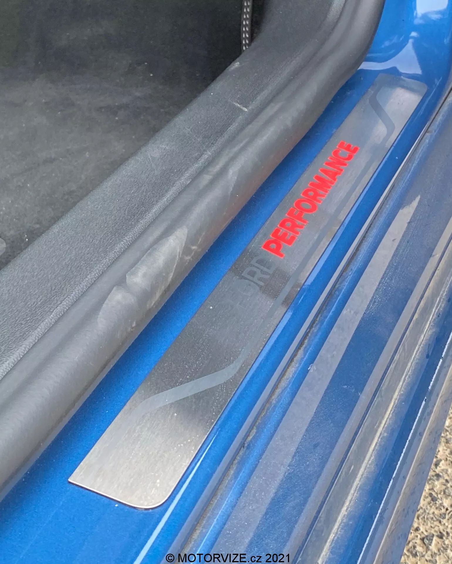 Fotografía en primer plano desde un ángulo alto muestra el área del umbral de la puerta del Ford Focus 2022. El enfoque está en la inscripción 'PERFORMANCE' en rojo, presentada en una inserción, lo que indica un modelo de alto rendimiento. Una placa protectora de aspecto metálico cepillado debajo protege el exterior pintado en azul del automóvil, que parece brillante y bien cuidado. La goma de sellado es visible a lo largo del borde superior.