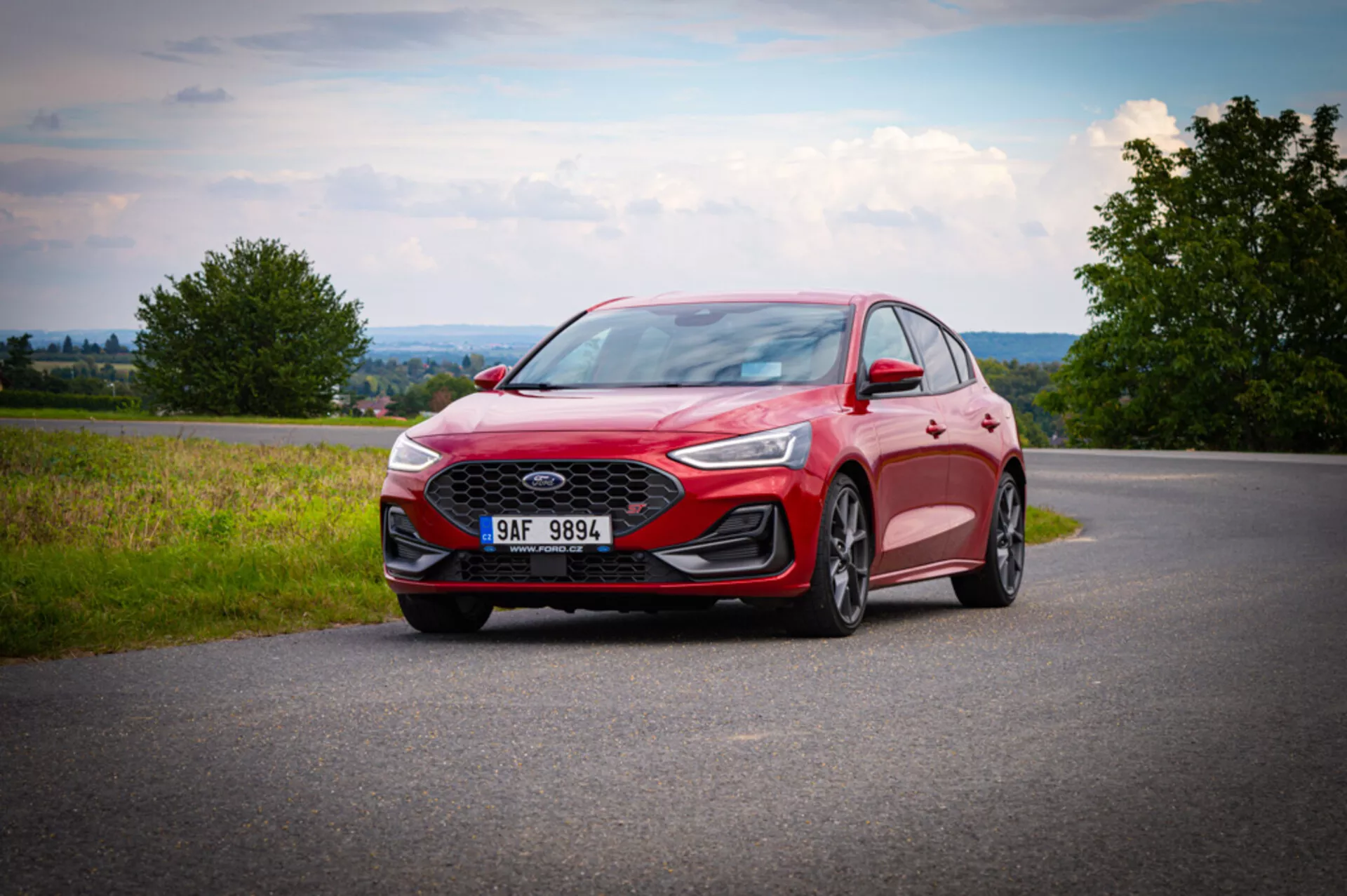 Ford Focus 2022 fotografiado desde un ángulo bajo frontal tres cuartos destacando la parrilla frontal con patrón de panal, faros LED delgados, ruedas de aleación con diseño de varios radios, capó contorneado, espejo lateral con señal de giro integrada, carrocería roja moderna con contornos aerodinámicos, posicionado en una carretera de asfalto con fondo campestre