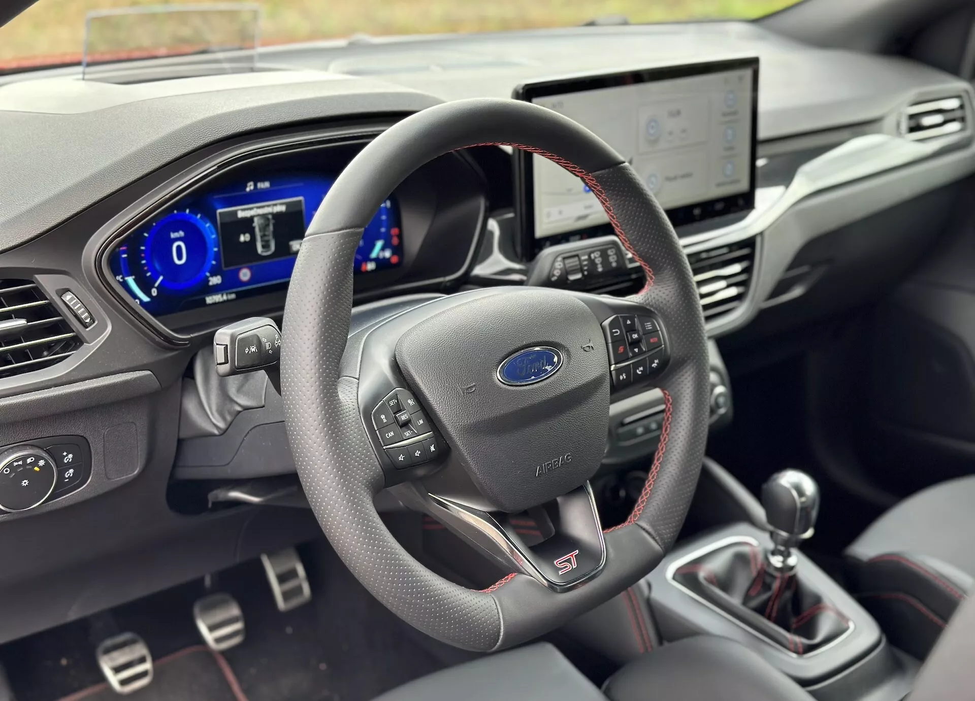 Interior del Ford Focus 2022 mostrando un volante forrado en cuero con controles integrados y la insignia 'ST', cuadro de instrumentos digital, sistema de infoentretenimiento táctil, botones de control climático, y palanca de cambios manual con materiales premium detallados y enfoque de profundidad de campo reducida