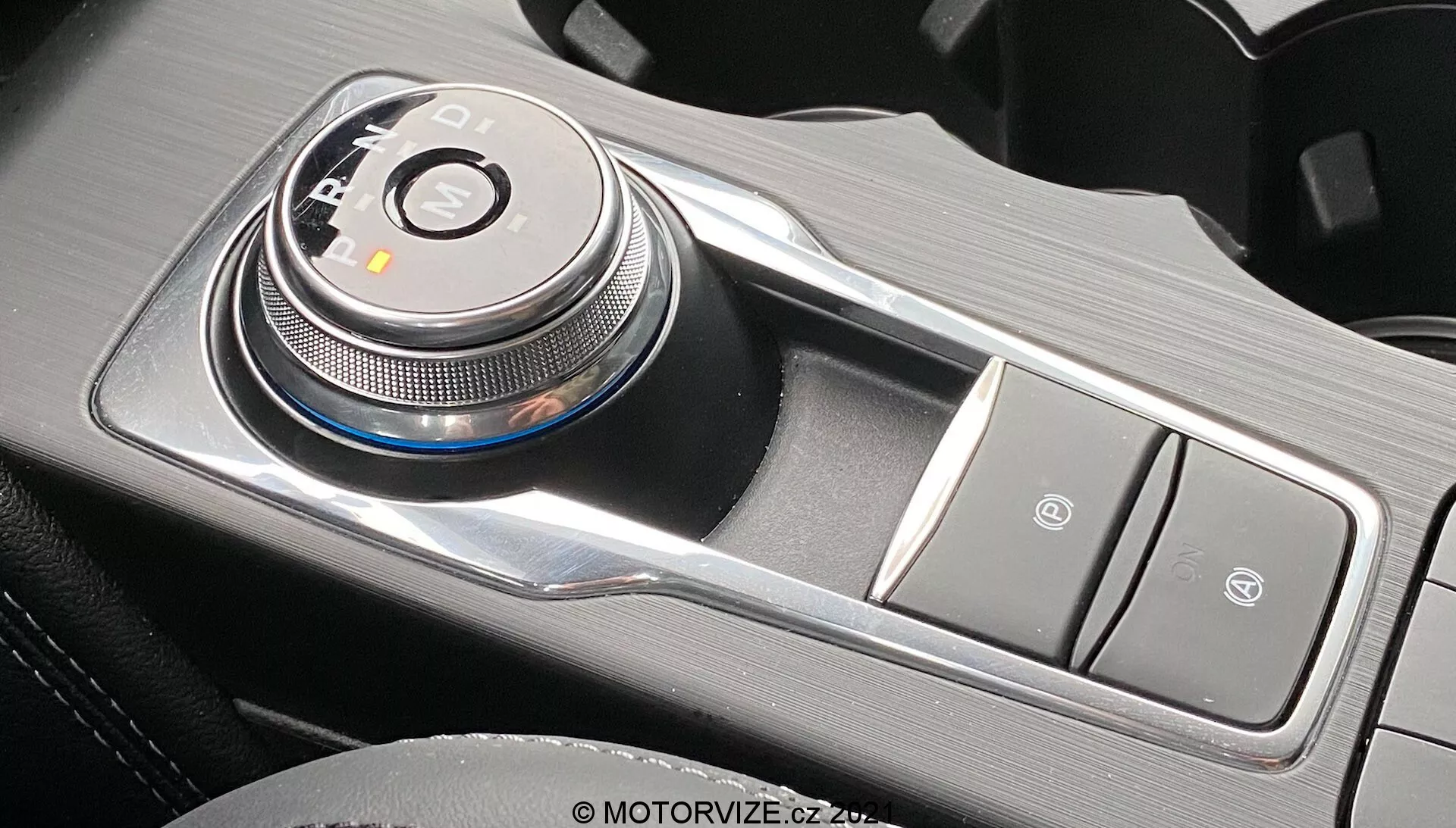 Imagen en primer plano del interior del Ford Focus 2022 que muestra un avanzado selector de cambios electrónico de dial giratorio situado hacia el lado del conductor en la consola central, en la posición 'P' de estacionamiento. El selector de cambios está rodeado por indicadores LED para P, R, N, D, posiblemente S o B, reflejando una transmisión automática con modos de deporte o frenado. A la derecha, botones dispuestos verticalmente, probablemente para el freno de estacionamiento electrónico y la función Auto-Hold. El acabado metálico y los materiales texturizados realzan la estética premium y moderna bajo la luz natural.