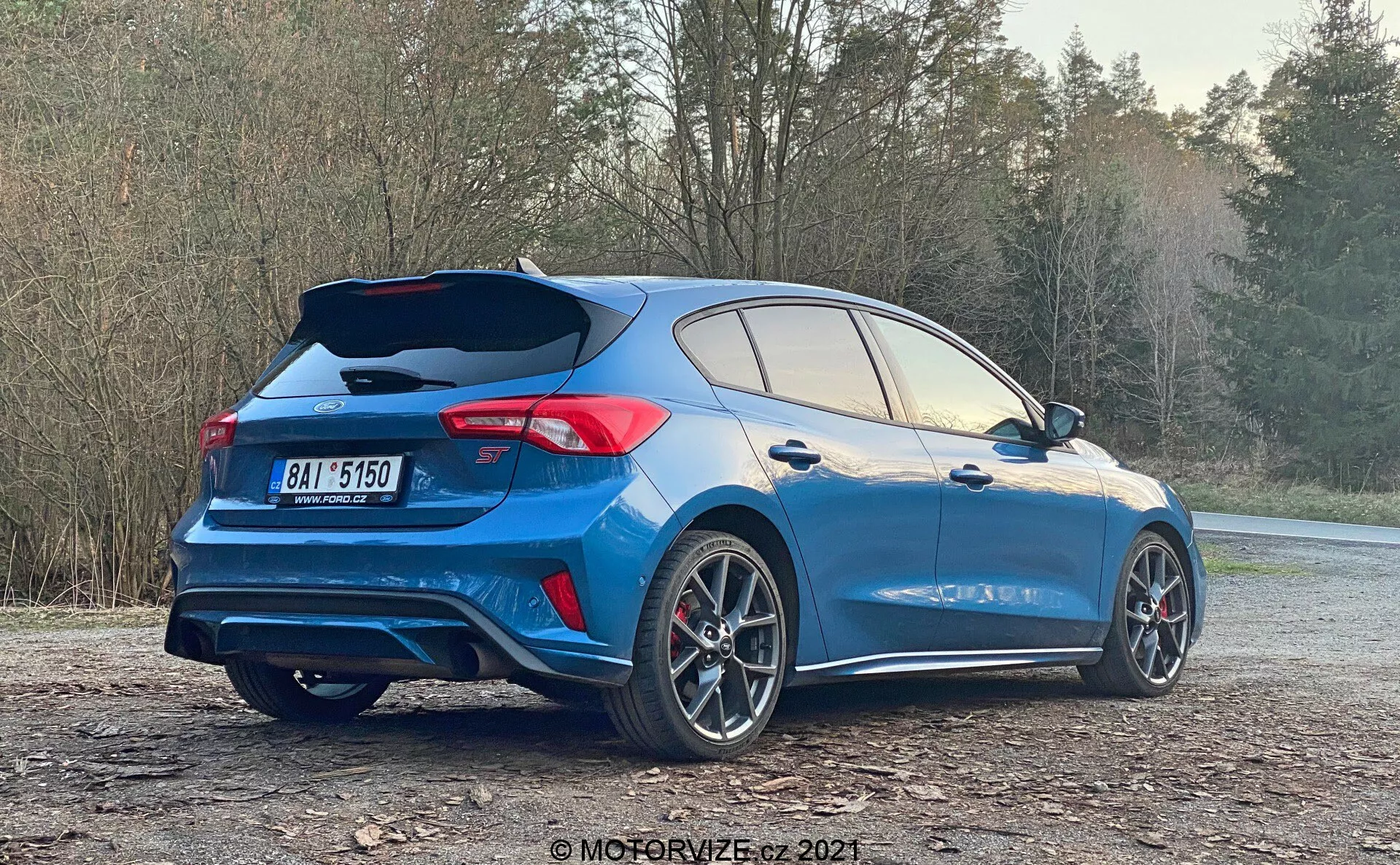 Vista trasera de tres cuartos de un coche hatchback moderno, destacando sus elementos deportivos como un alerón en el techo, esculpido aerodinámico y tubos de escape dobles. El coche es azul con detalles en negro o gris oscuro, grandes ruedas de aleación multirradiales con pinzas de freno visibles y luces traseras LED. La puerta trasera abierta revela un amplio espacio de carga, sugiriendo fácil accesibilidad. Aparcado en un área de gravilla con árboles en el fondo, esta imagen enfatiza el diseño y las características del vehículo adecuadas para un estilo de vida activo.