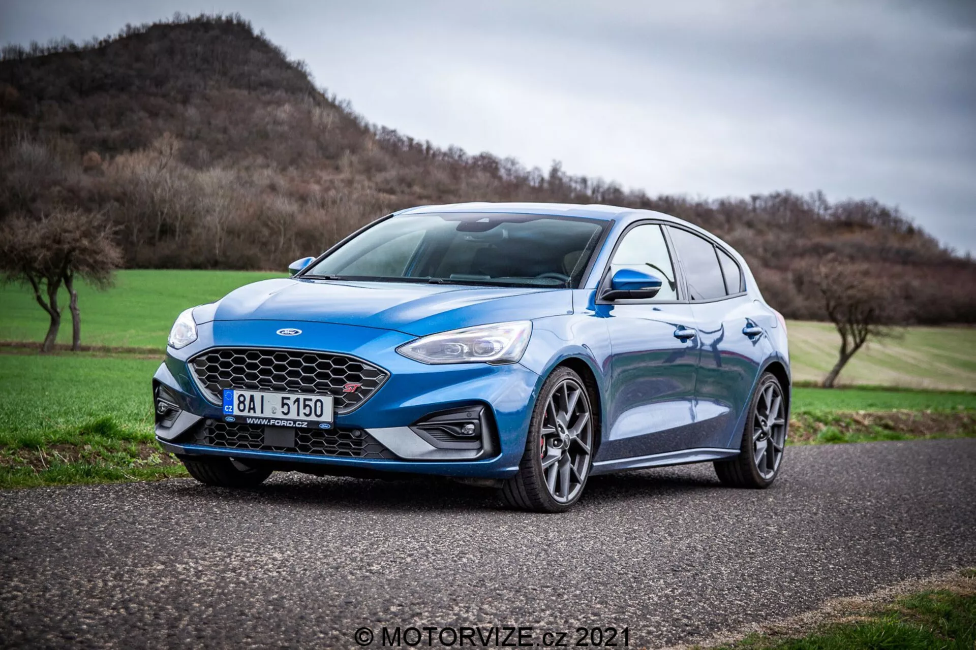 Ford Focus 2022 azul capturado desde un ángulo bajo con una vista de tres cuartos frontal, mostrando su diseño aerodinámico, moderno y deportivo. Las características destacadas incluyen la parrilla en forma de panal con un emblema central, elegantes faros LED, pliegues dinámicos en el capó, grandes ruedas de aleación de múltiples radios, neumáticos de perfil bajo, espejos laterales con intermitentes integrados y un indicador lateral de lente clara en el guardabarros delantero. El coche está enfocado con nitidez contra un fondo suavemente difuminado de un paisaje montañoso y un cielo nublado, sugiriendo que es un hatchback deportivo centrado en el rendimiento con opciones avanzadas de iluminación automotriz y características de seguridad.