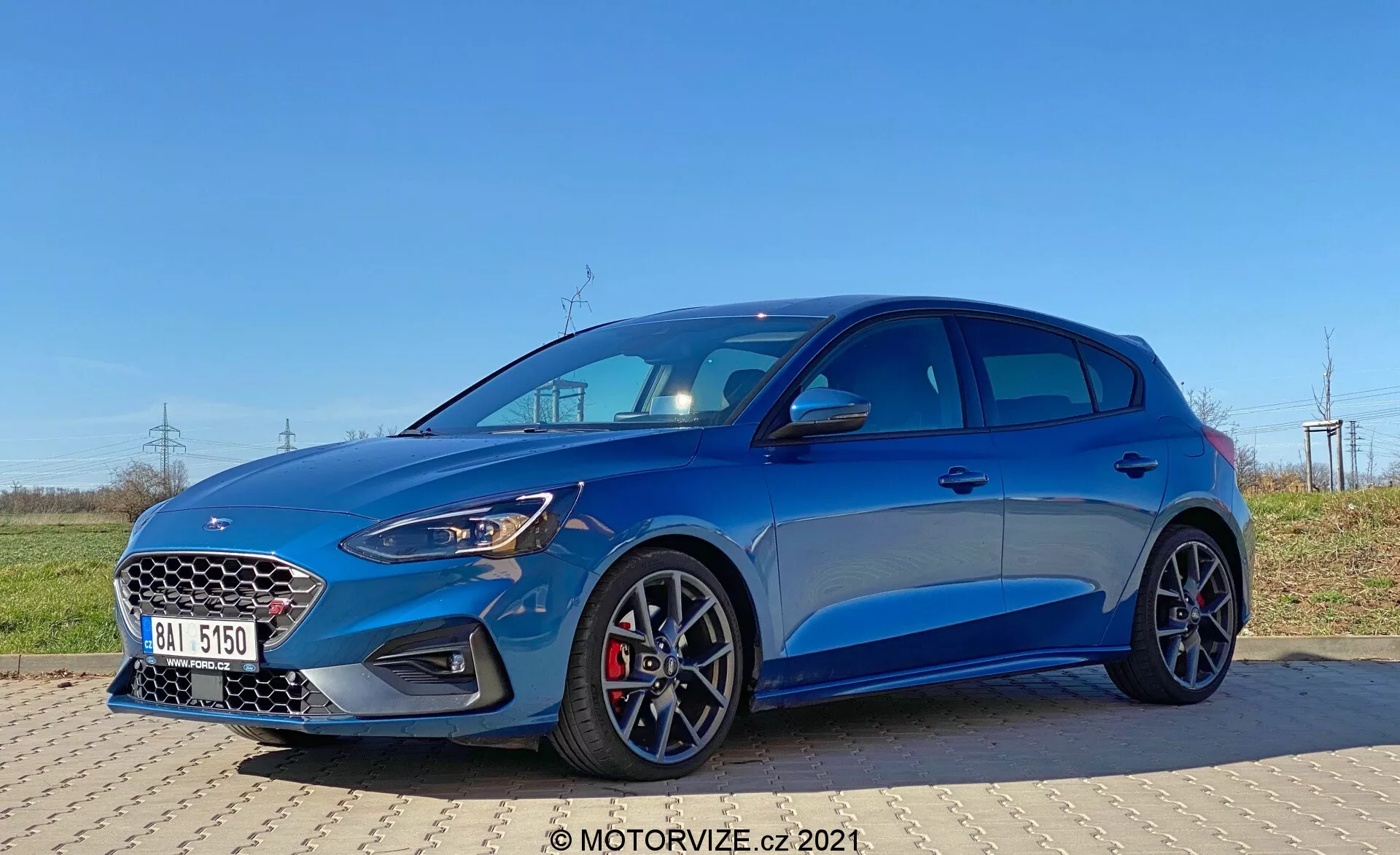 Imagen de un Ford Focus 2022 capturado desde un ángulo frontal de tres cuartos, mostrando la parrilla delantera con patrón de panal, faros LED, diseño moderno del parachoques con tomas de aire más grandes y un capó elegante. El hatchback metálico azul muestra una silueta deportiva, pinzas de freno rojas, ruedas multirradio, líneas de carácter afiladas, arcos de ruedas prominentes y espejos laterales con señales de giro integradas, todo resaltado bajo condiciones de luz diurna clara.