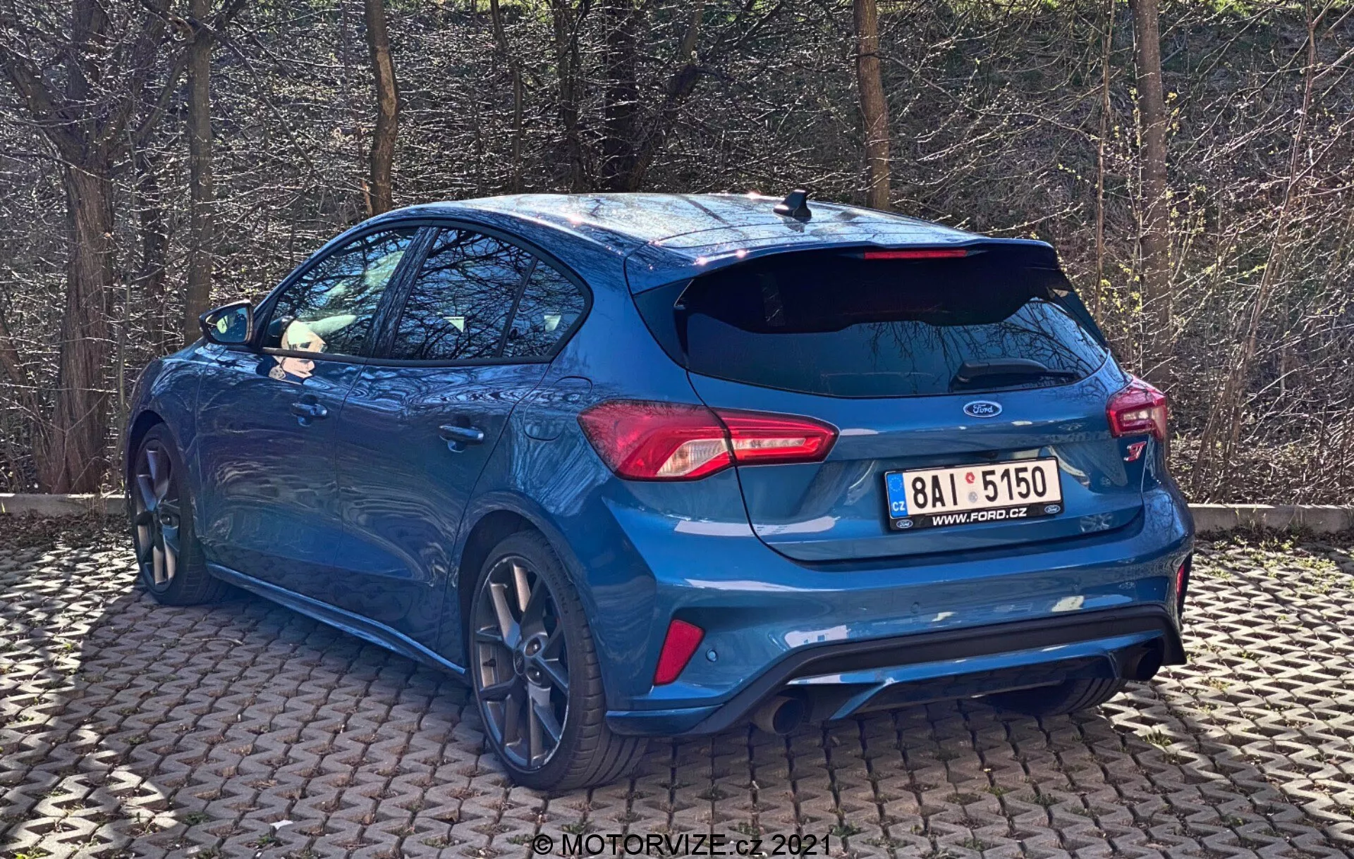 Ford Focus 2022 azul capturado desde un ángulo trasero de tres cuartos, destacando el diseño deportivo del hatchback con alerón de techo, grandes llantas de aleación con bordes de corte de diamante, luces traseras LED elegantes, parachoques trasero prominente con sensores integrados, doble tubo de escape, cámara de visión trasera, acabado de pintura metálica brillante y vidrios polarizados, estacionado sobre una superficie de ladrillo con luz natural y árboles de fondo.
