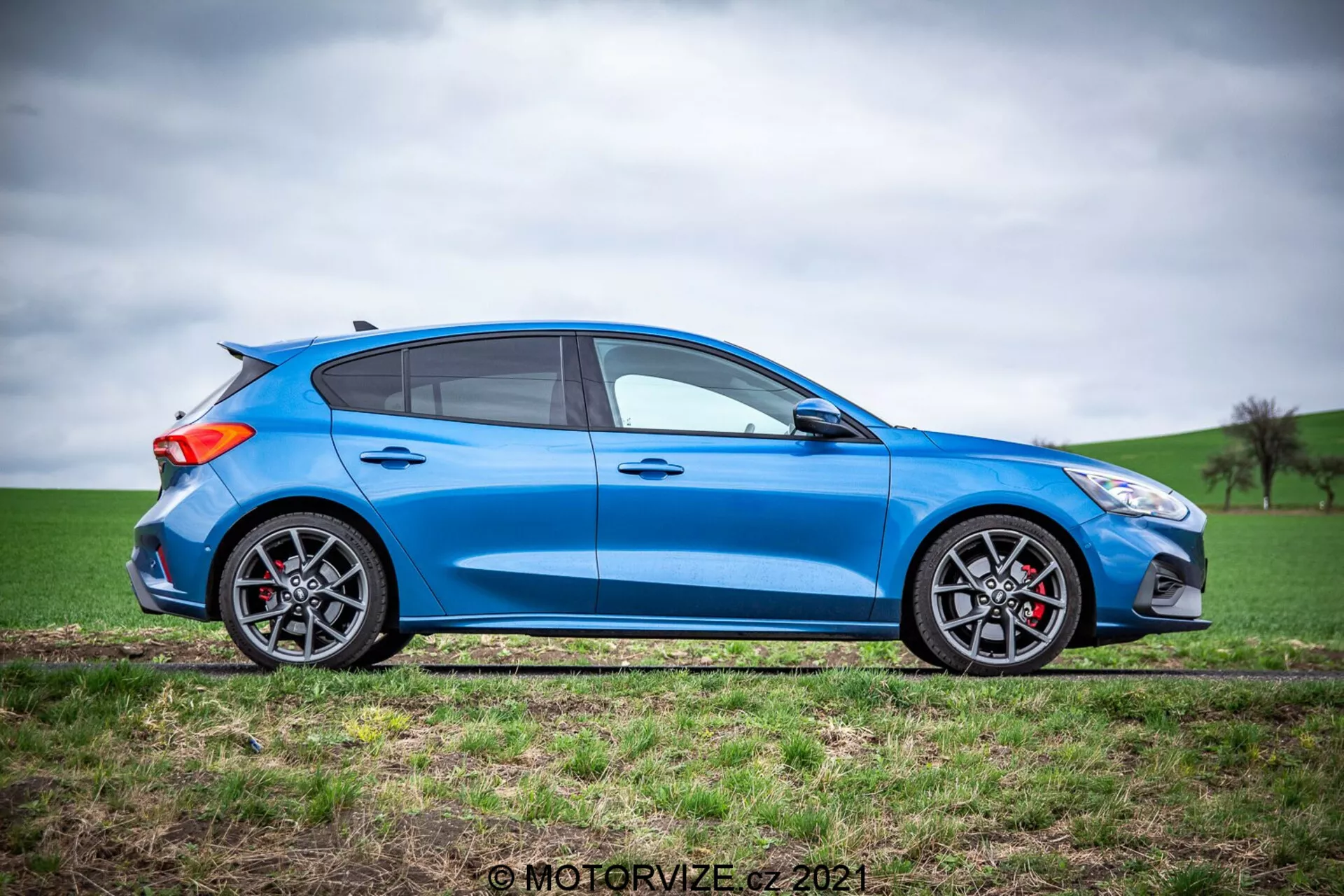 Una vista de perfil lateral completo de un hatchback Ford Focus 2022 de color azul brillante a la luz natural del día con una silueta aerodinámica y deportiva, cuatro puertas, ventanas con marcos negros y pilar B negro, espejos laterales negros, pinzas de freno rojas a través de ruedas de aleación con llantas de perfil bajo y un pequeño alerón trasero sobre una superficie relativamente plana adyacente a un campo con hierba verde y un cielo nublado.
