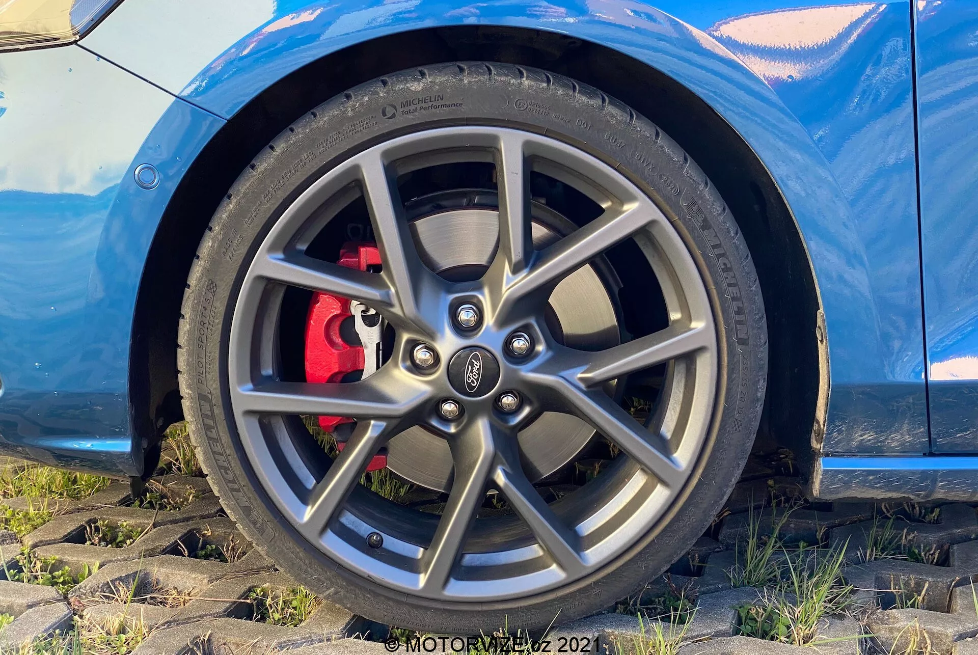 Vista detallada del conjunto de rueda y neumático del Ford Focus 2022 desde una perspectiva baja y ligeramente angulada. La imagen enfatiza el borde de aleación de múltiples radios, el neumático de alto rendimiento de perfil bajo y la pinza de freno roja detrás del borde, lo que sugiere un diseño deportivo. En la parte superior de la imagen se puede ver una porción de la carrocería del automóvil en color azul metálico, contribuyendo a la apariencia elegante y dinámica.