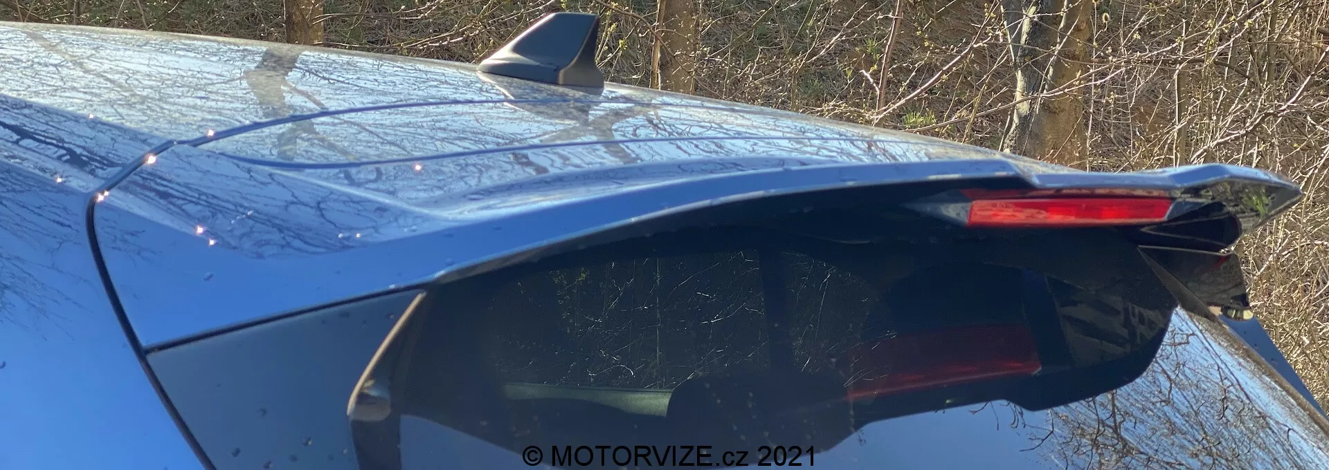 Vista en ángulo trasero cercano del Ford Focus azul 2022 que muestra la parte superior trasera, con un spoiler trasero integrado elegante con una tercera luz de freno al final de la línea del techo, sugiriendo estabilidad y diseño deportivo. La imagen también muestra una antena de aleta de tiburón en el techo, insinuando sistemas de comunicación modernos. El parabrisas trasero inclinado y afilado realza el aspecto deportivo, con gotas de agua en la superficie que indican condiciones exteriores. La calidad reflectante de la pintura sugiere una buena iluminación y un acabado limpio.