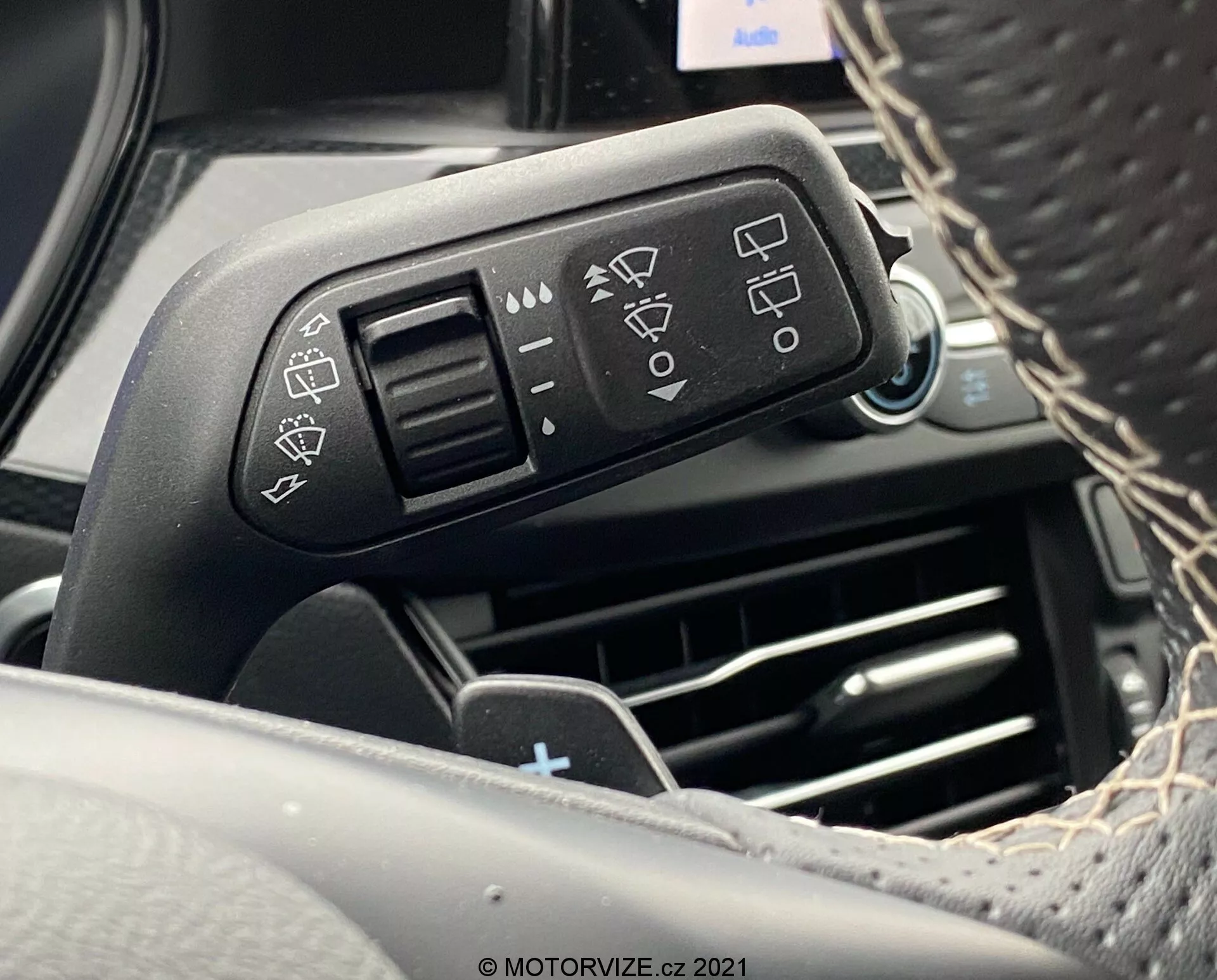 Vista en primer plano de la palanca de control montada en la columna de dirección del Ford Focus 2022 desde la perspectiva del conductor. La imagen se enfoca en la palanca de control del lado izquierdo con íconos grabados para configuraciones de faros, indicadores de señal de giro y limpiaparabrisas. Los reflejos brillantes en la palanca resaltan la calidad del material. El fondo cuenta con características como ventilaciones de aire acondicionado y elementos del tablero, ligeramente desenfocados para enfatizar los detalles de la palanca.