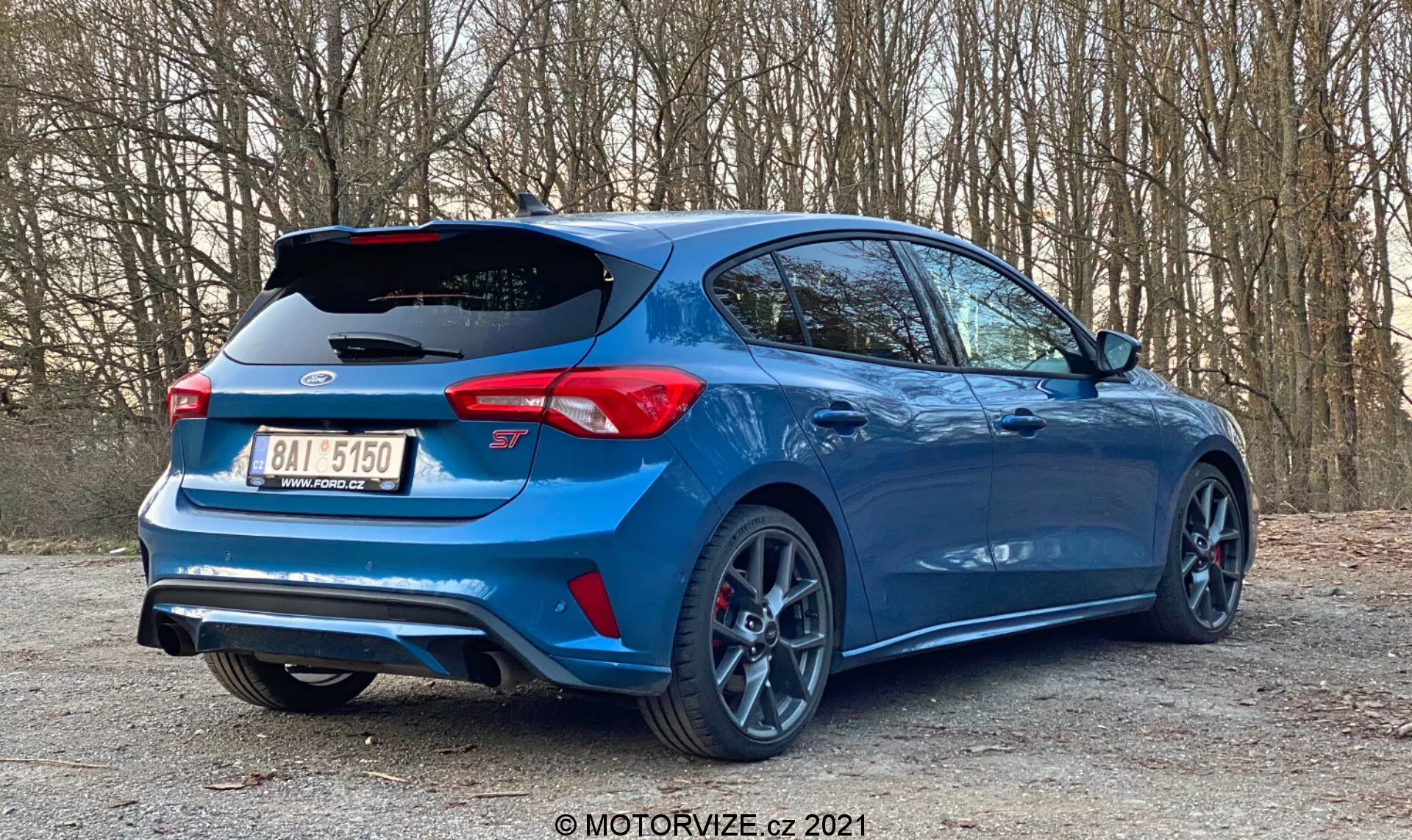 Un hatchback moderno Ford Focus 2022 aparcado en una área apartada junto a una carretera con árboles en el fondo, visto desde un ángulo bajo tres cuartos trasero. Se muestran las características traseras del coche, el perfil lateral, las ruedas, un alerón aerodinámico, dobles salidas de escape, difusor trasero, luces traseras LED, la insignia del modelo, un parachoques trasero distintivo, llantas deportivas de múltiples radios con neumáticos de perfil bajo, una pinza de freno roja de alto rendimiento, la pintura metálica azul y ventanas tintadas, destacando elementos de diseño automotriz, rendimiento y tecnología de seguridad.