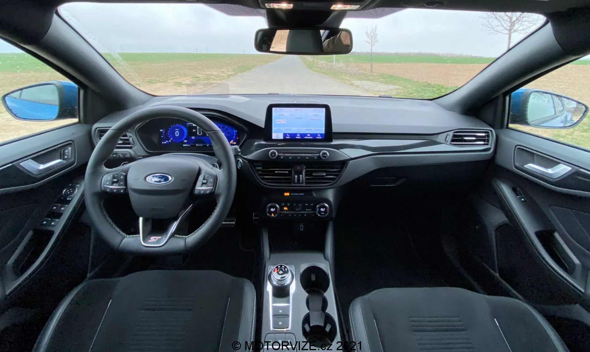Vista interior del Ford Focus 2022 desde el área de los asientos traseros, destacando el moderno volante multifuncional con controles integrados, panel de instrumentos totalmente digital, sistema de infoentretenimiento con pantalla táctil, botones de control de clima brillantes, palanca de cambios giratoria, asientos delanteros contorneados tapizados en material oscuro y paneles de puerta a juego con manijas integradas y superficies de control para ventanas y espejos.