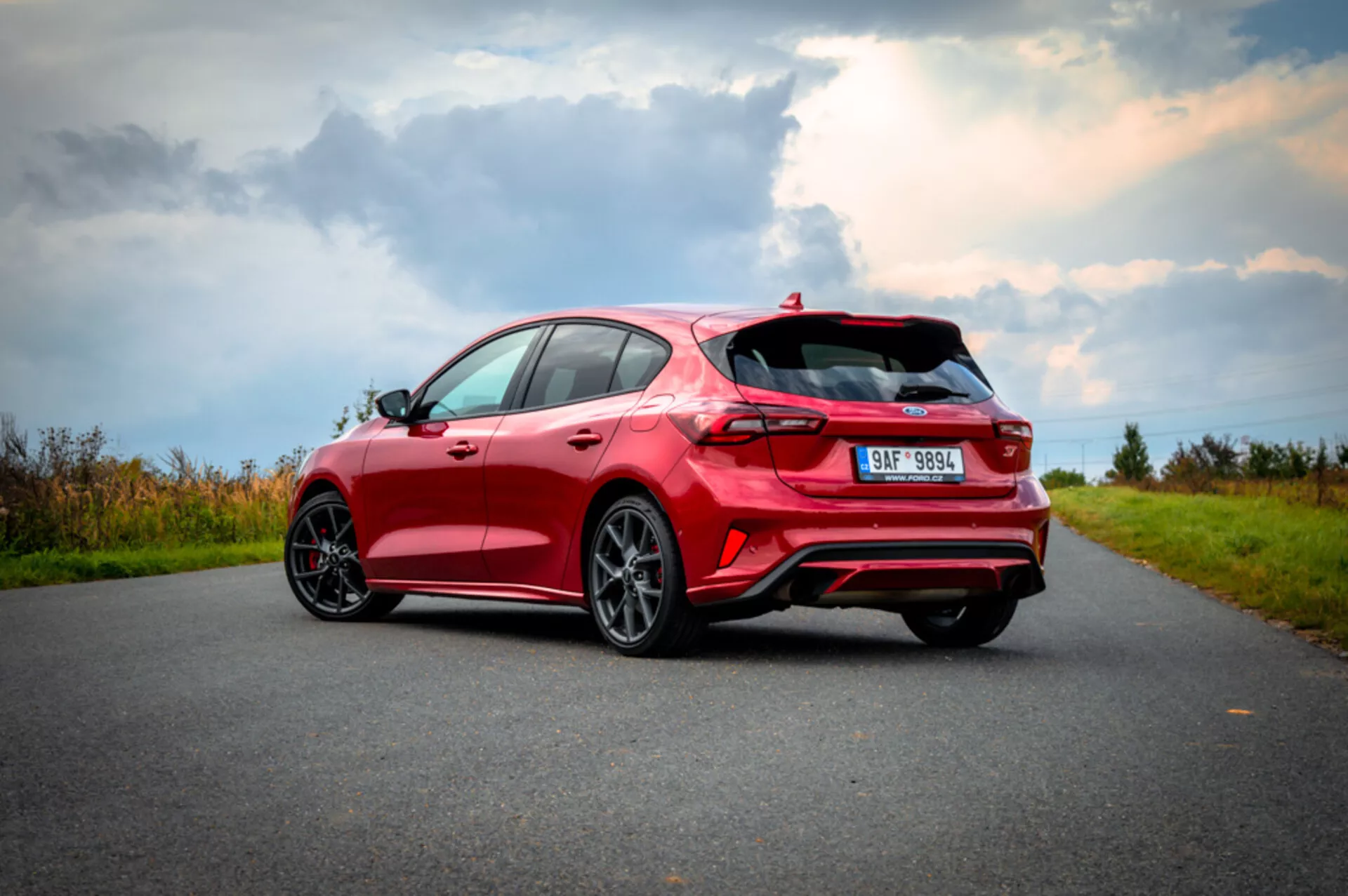 Vista trasera tres cuartos del lado derecho del Ford Focus hatchback rojo 2022, destacando diseño aerodinámico con alerón, luces traseras LED, sistema de escape doble, ruedas de acabado oscuro multiradio y neumáticos de perfil bajo, en una carretera de asfalto con paisaje natural de fondo y cielo parcialmente nublado